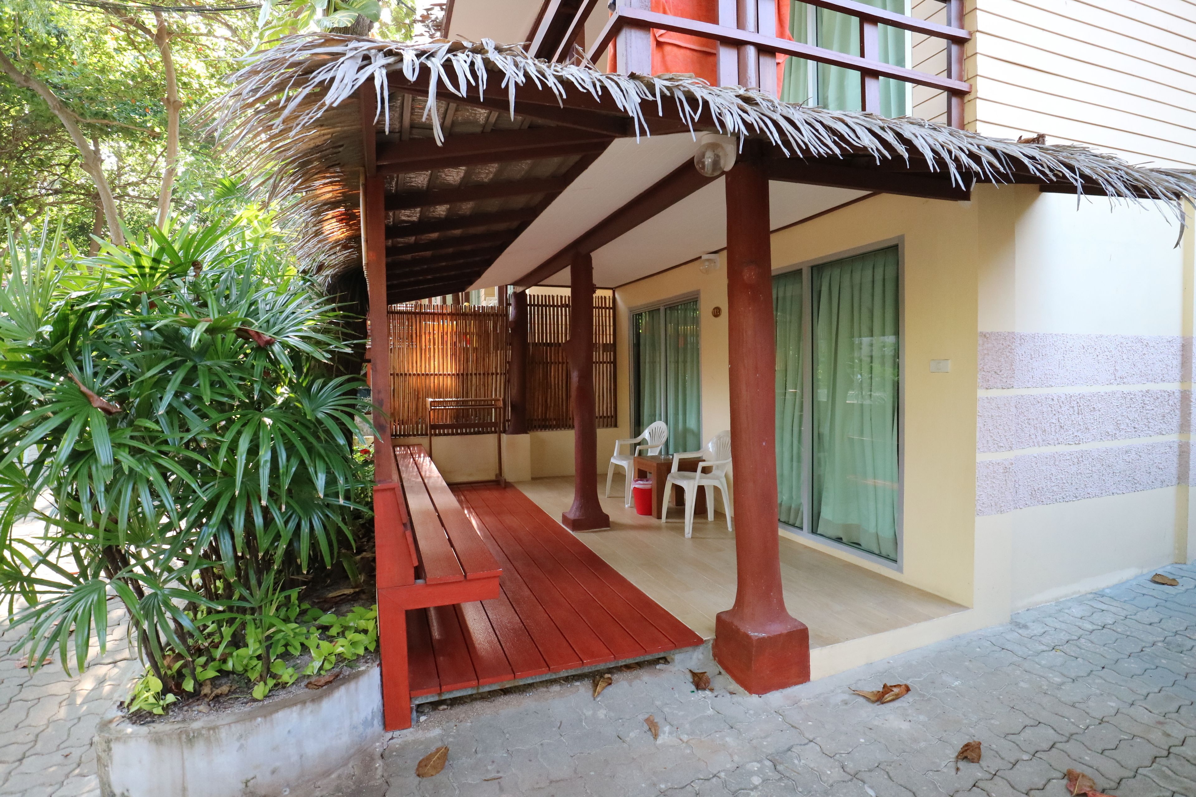 A covered outdoor seating area with a wooden bench, a concrete planter with green plants, and a building entrance with a glass door and a window.