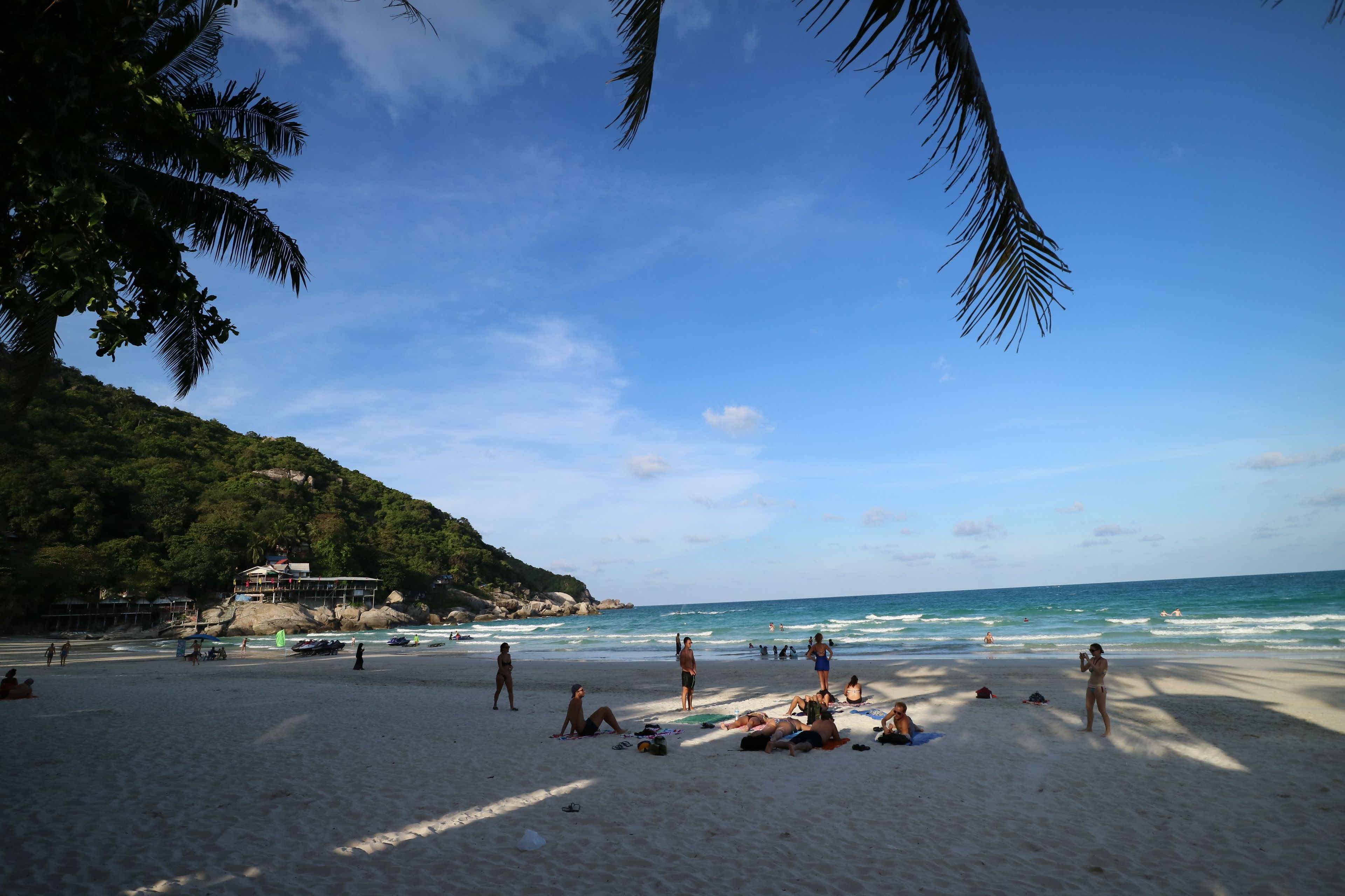 Ein Strand mit badenden und sonnenbäckernden Personen, ein Gebäude auf einem Hügel und strahlend blauem Himmel.