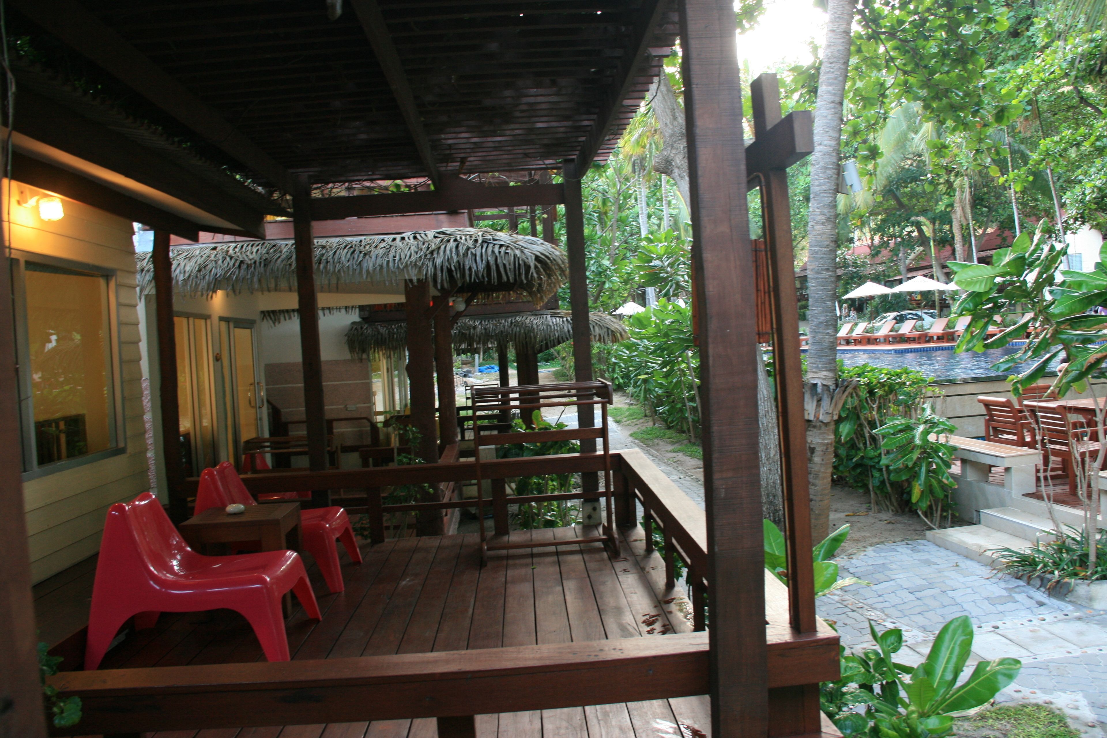 A wooden deck with red plastic chairs and tables, surrounded by thatched umbrellas and lush greenery.