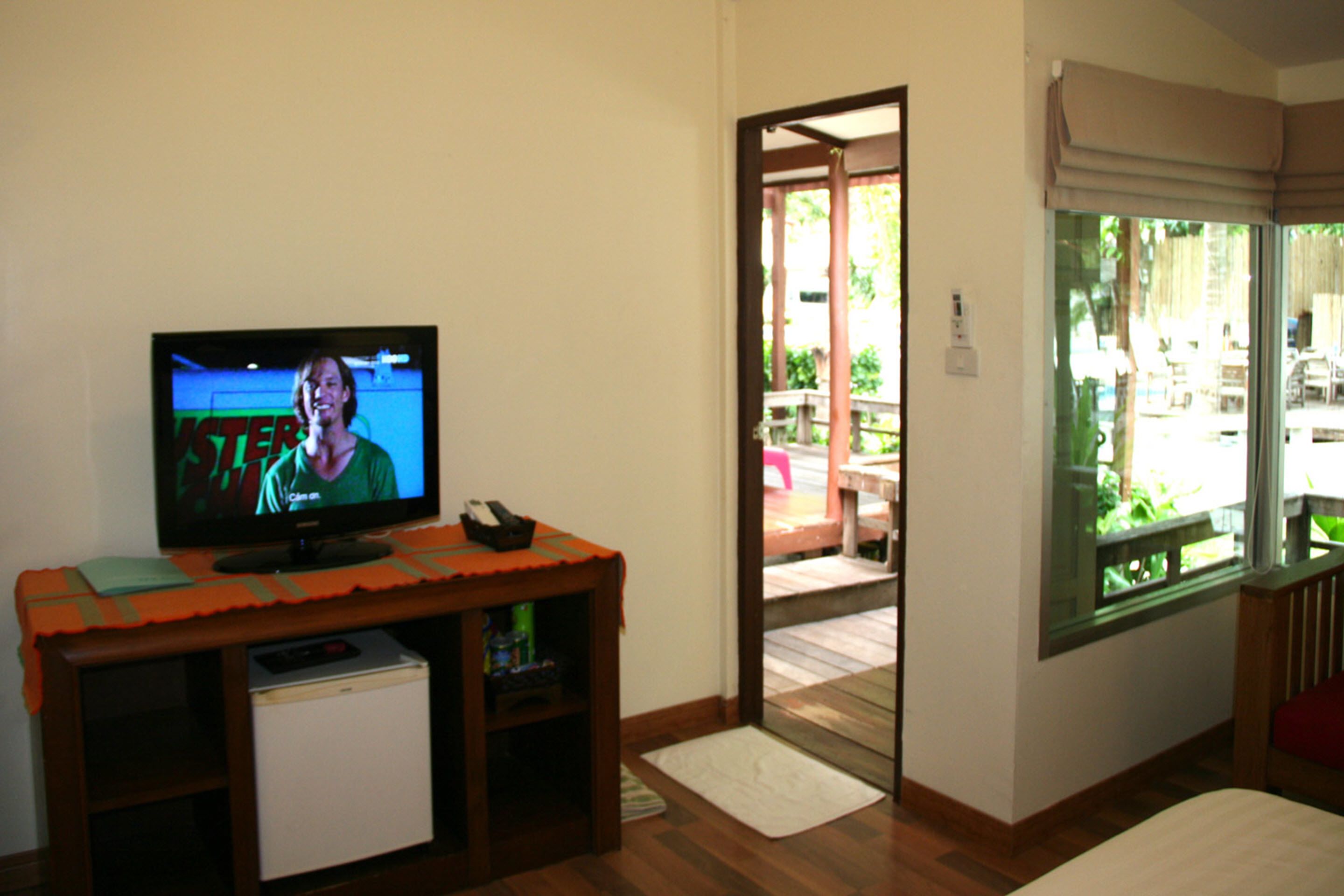 Una habitación con una televisión sobre un soporte de madera, un refrigerador pequeño y una puerta corrediza de vidrio que da a un área exterior.