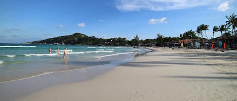 A beach with clear skies, people walking along the shore, and buildings in the background.