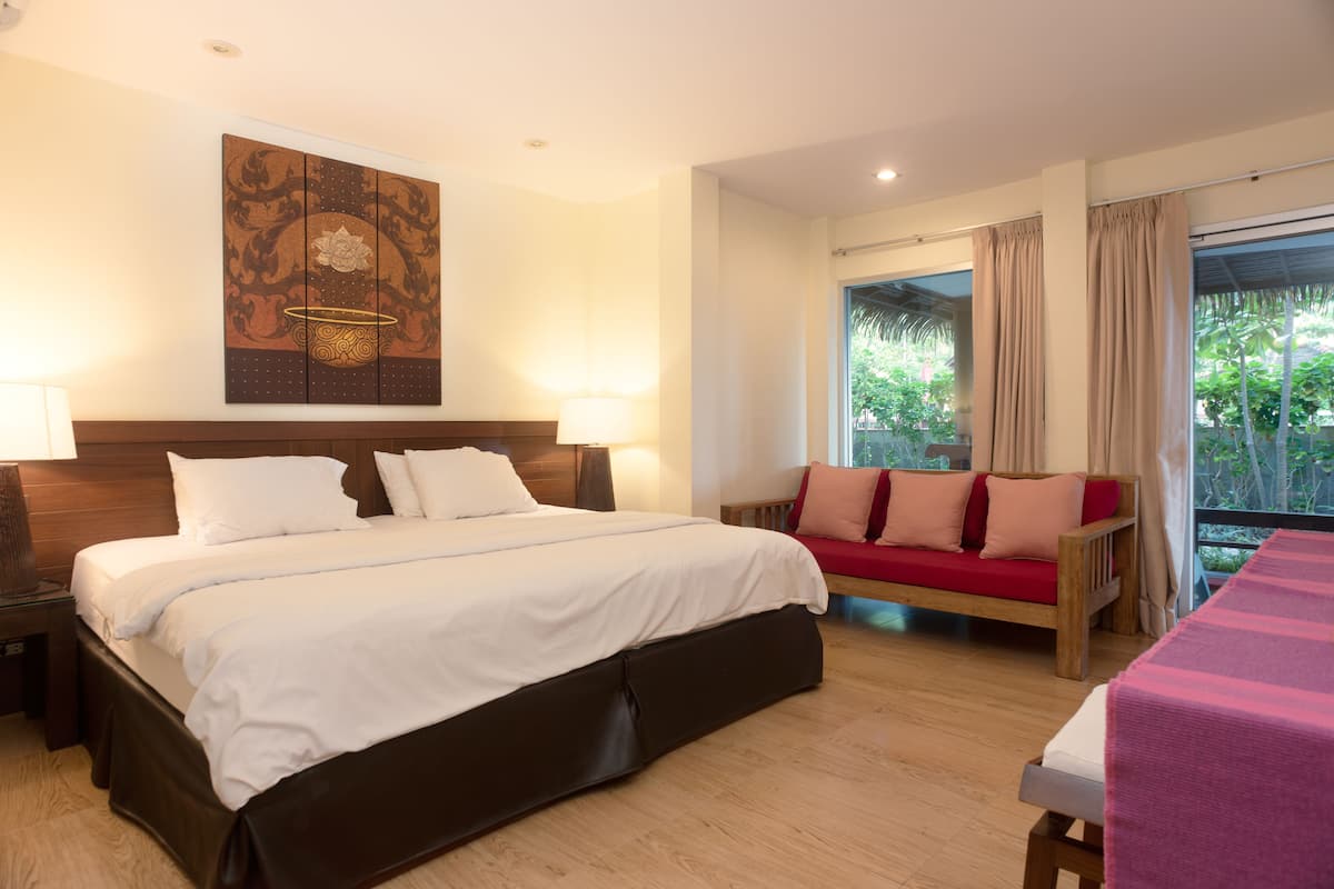 A hotel room with a large bed, a wooden headboard, a sofa with cushions, a table, and a view of greenery through the window.