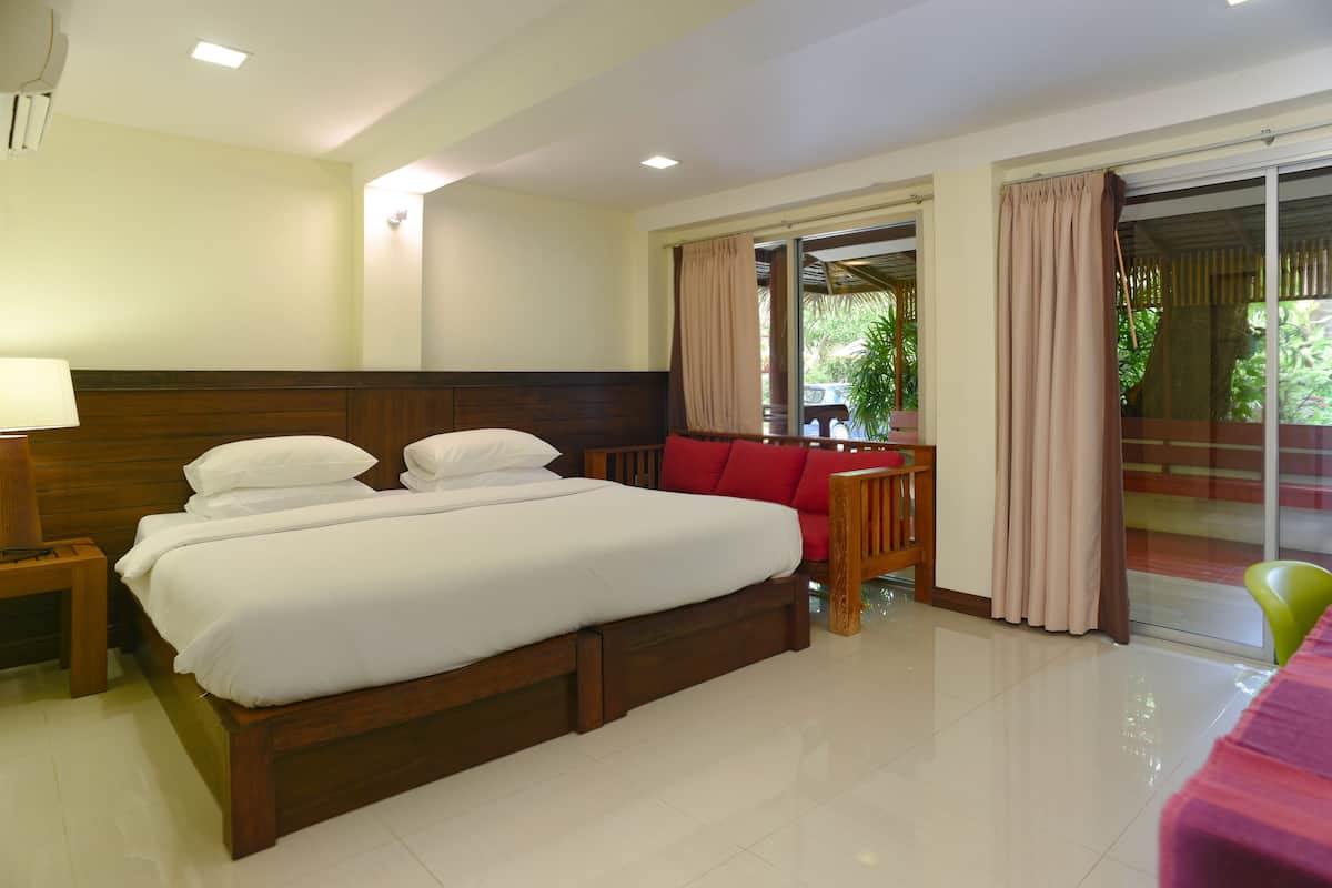 A hotel room with a large bed, a red sofa, a small table, and a sliding glass door leading to a balcony.