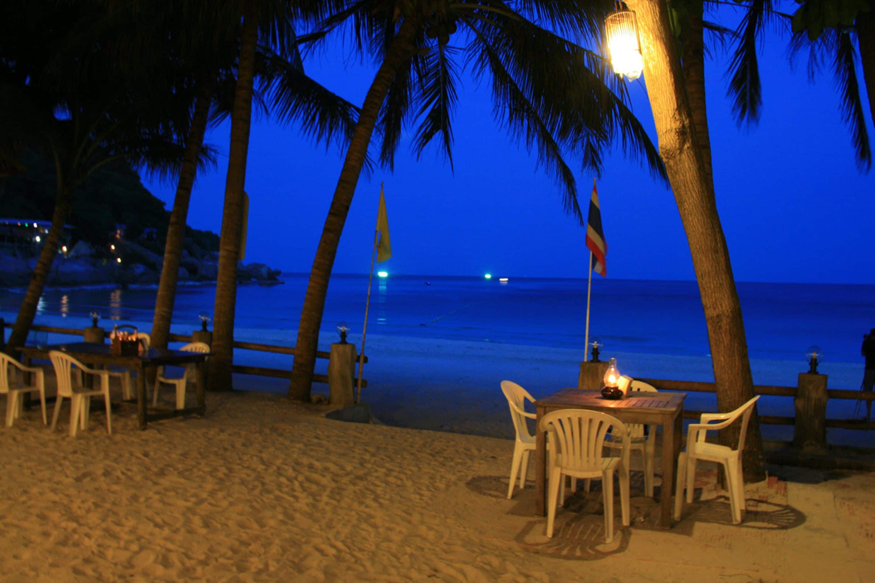 Un restaurante frente a la playa con mesas al aire libre, palmeras y vista al océano al atardecer.