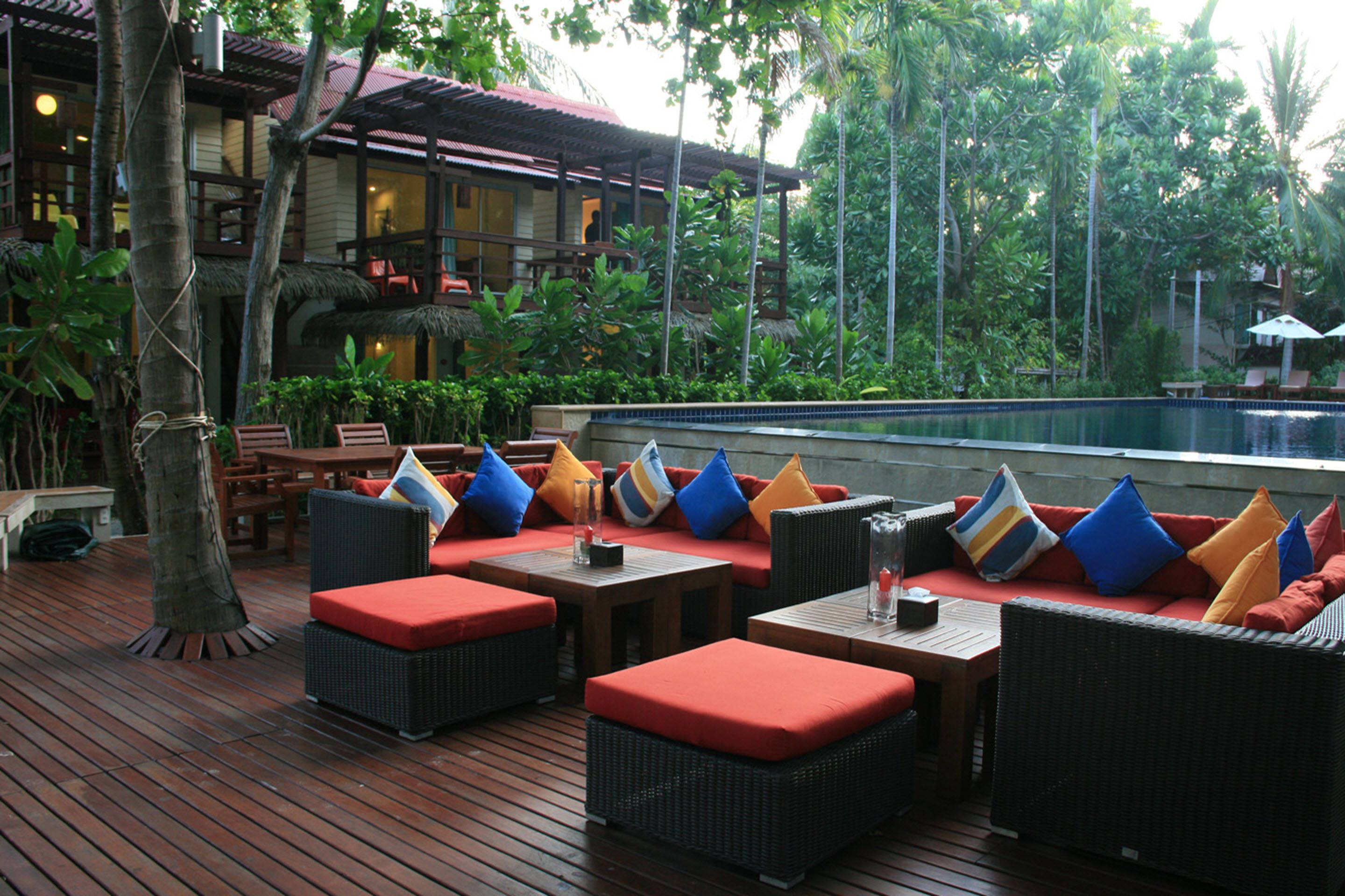 Un espacio lounge al aire libre con muebles de mimbre y cojines rojos, con vistas a una piscina y abundante follaje.
