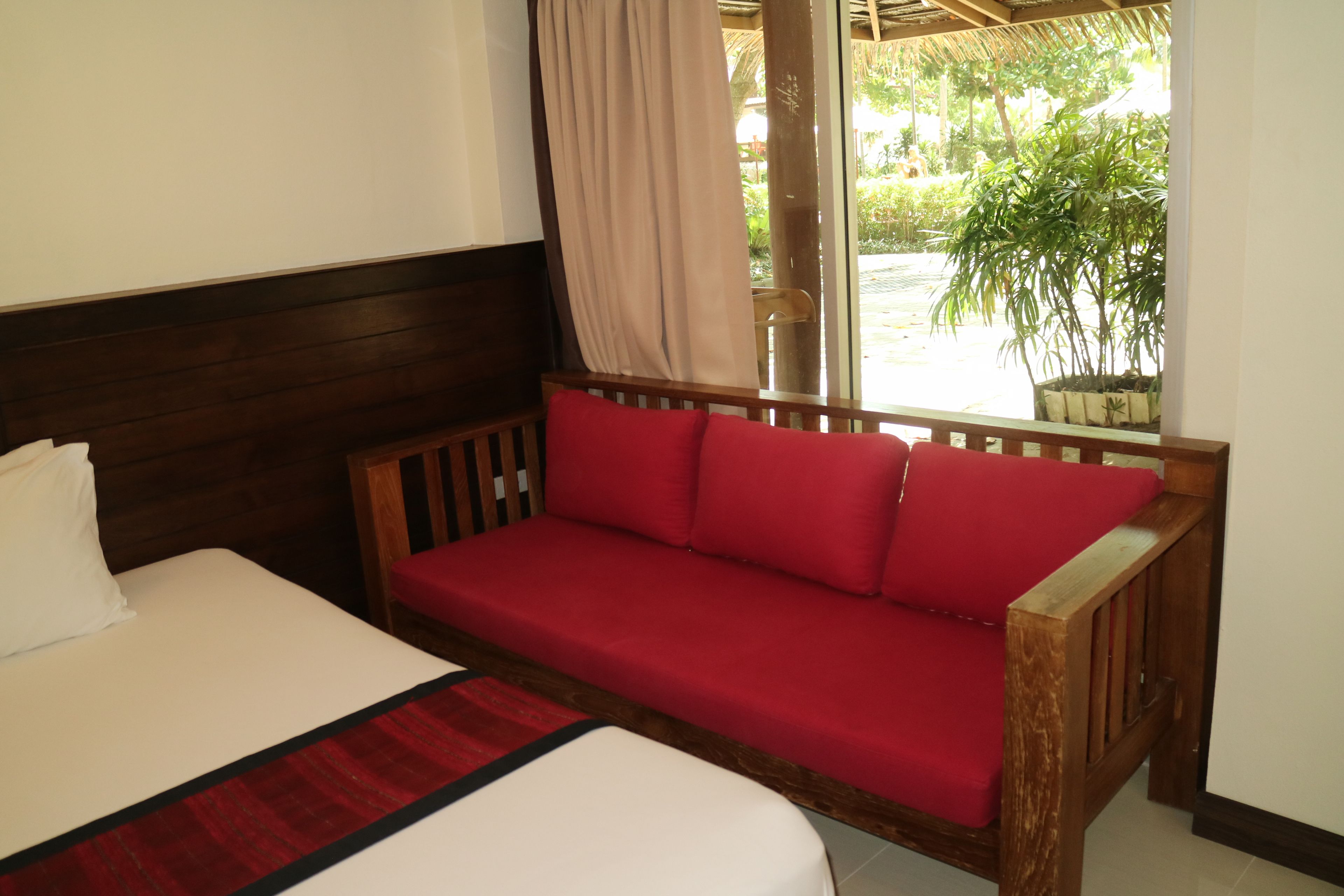 A room with a bed, a wooden sofa with red cushions, a window with a view of greenery, and a curtain.