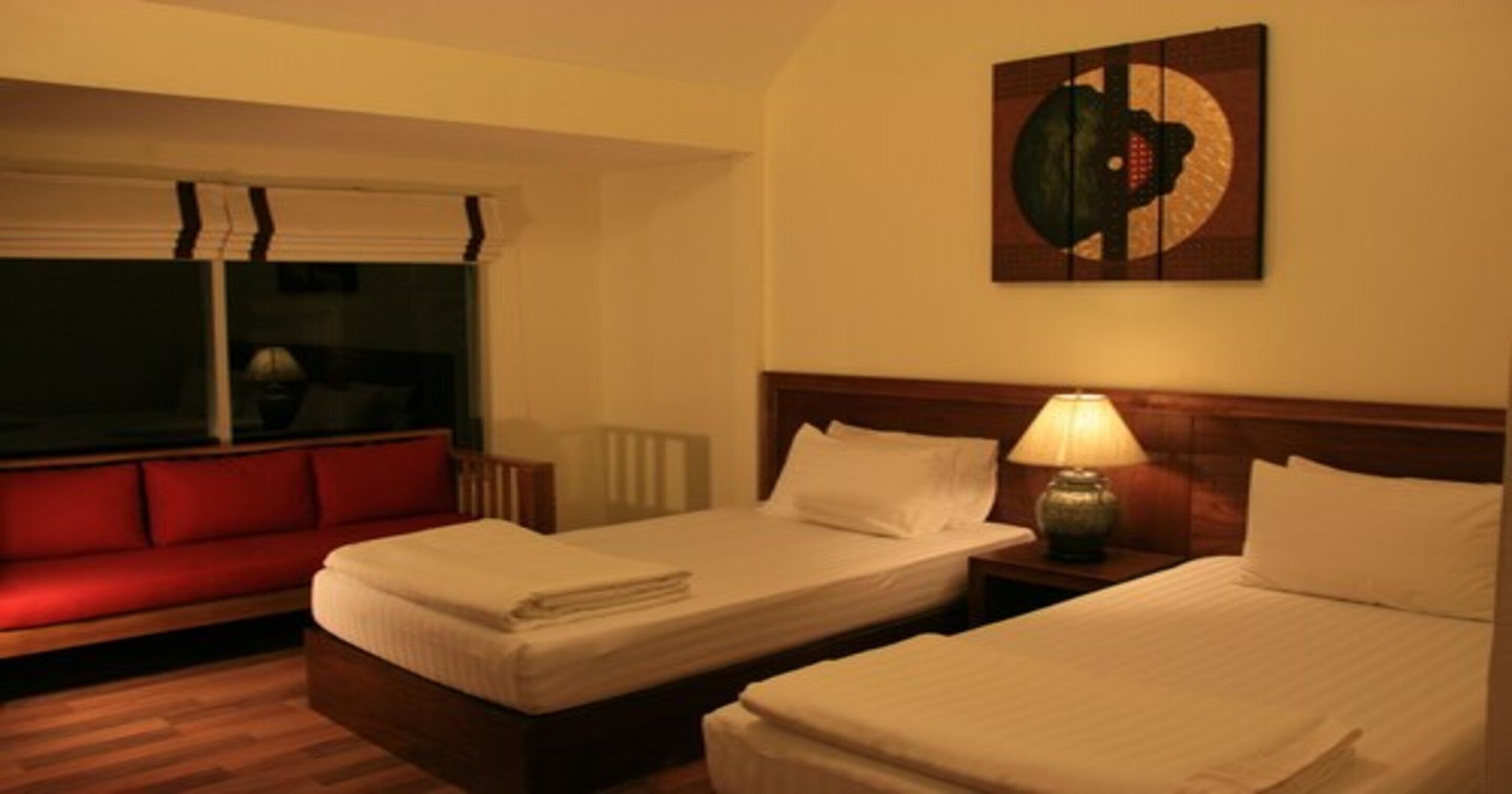 Habitación de hotel con dos camas, un sofá rojo, una ventana con persianas y una obra de arte colgada en la pared.