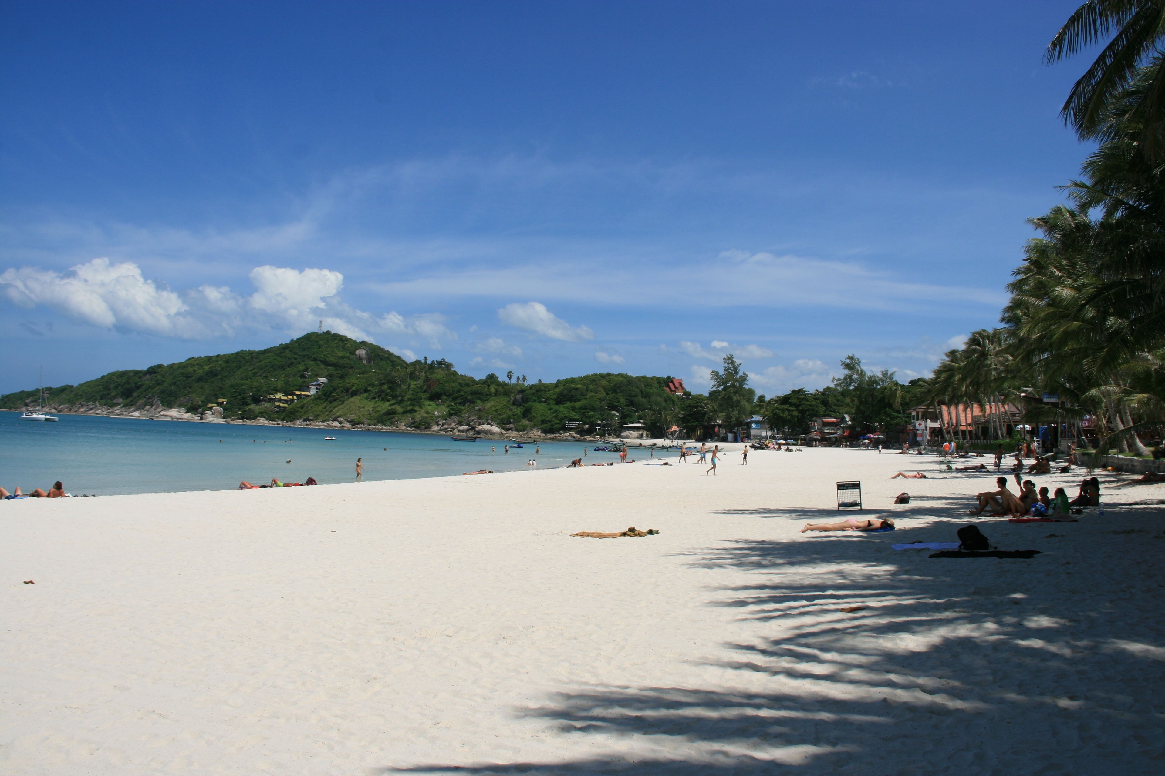 A beach with white sand, clear blue water, and a hill in the background.