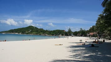 A beach with white sand, clear blue water, and a hill in the background.