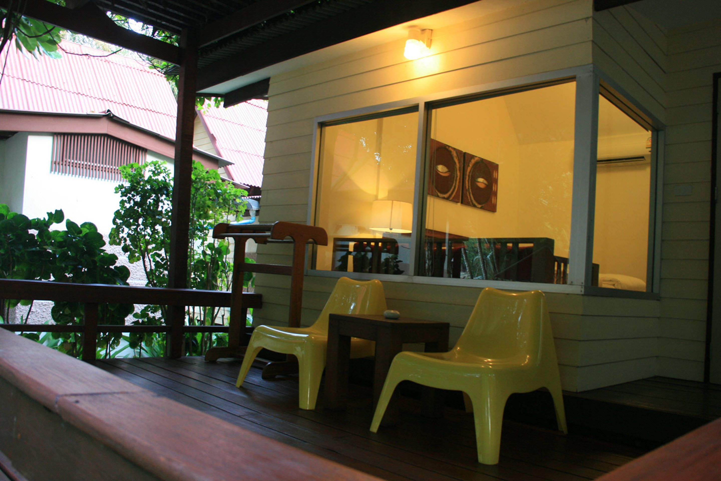 A covered outdoor seating area with wooden benches and yellow plastic chairs, a wooden table, and a window with a decorative item.