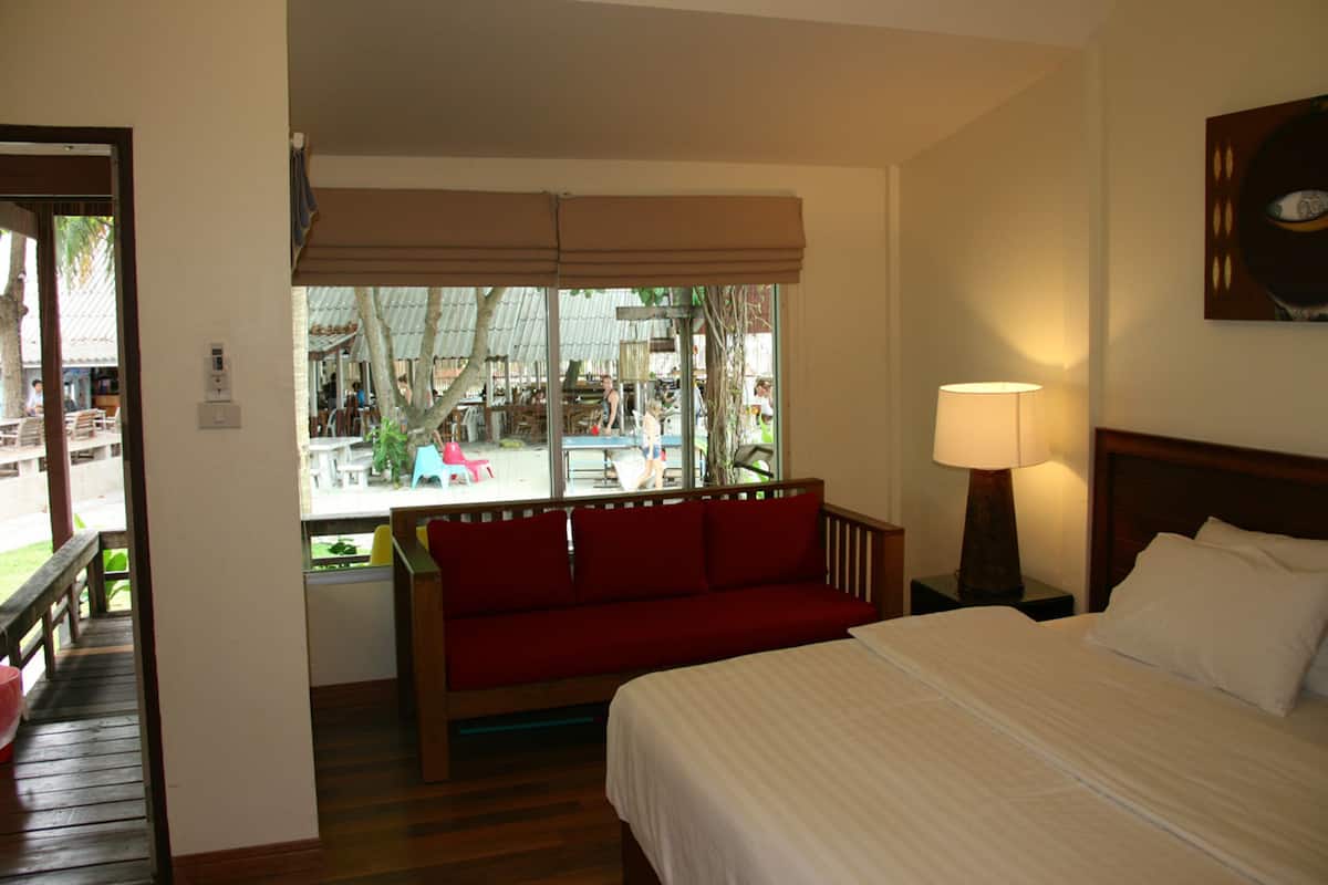 A hotel room with a bed, a red sofa, a window with a view of the outside, and a lamp on the nightstand.