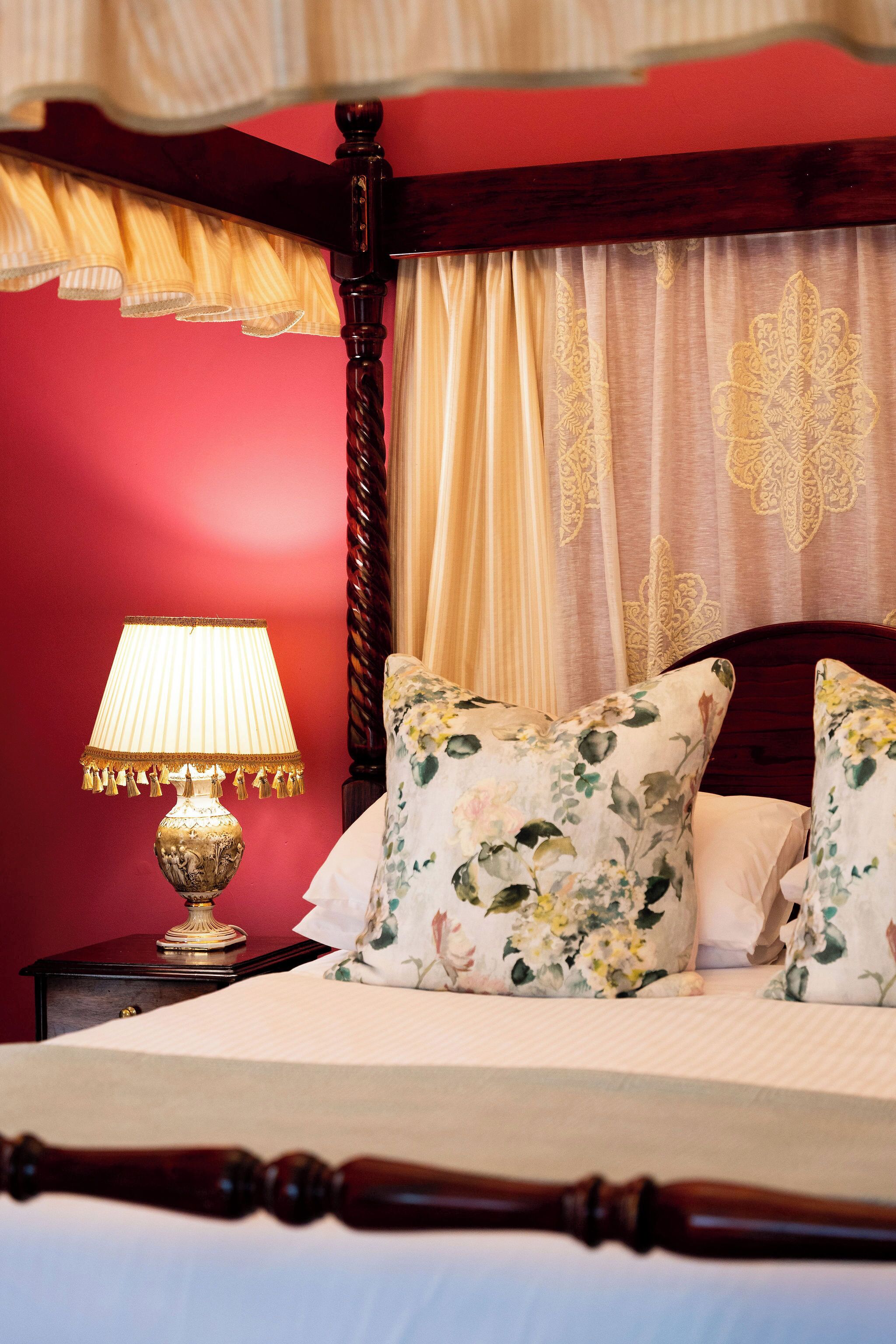 A bed with a canopy, a nightstand with a lamp, floral pillows, and a red wall.