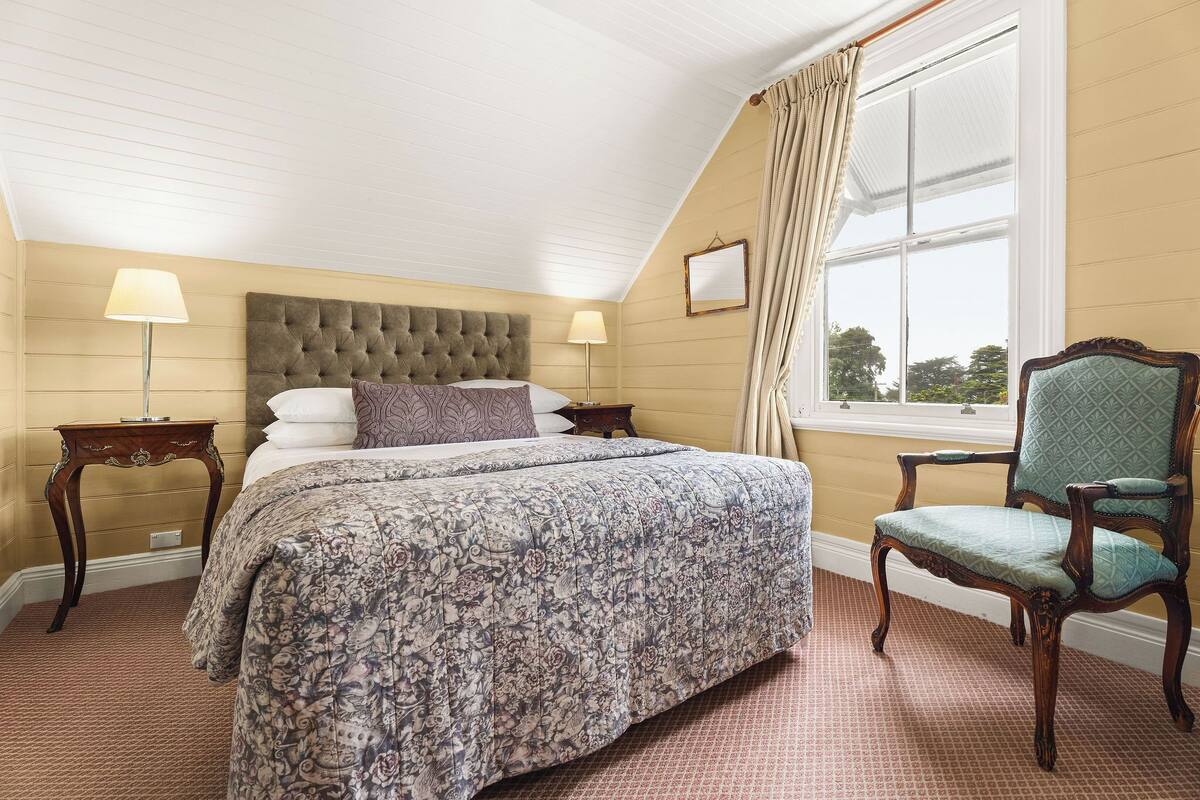 Une chambre avec un grand lit, deux tables de chevet, une chaise, une fenêtre avec des rideaux et une vue sur les arbres à l’extérieur.