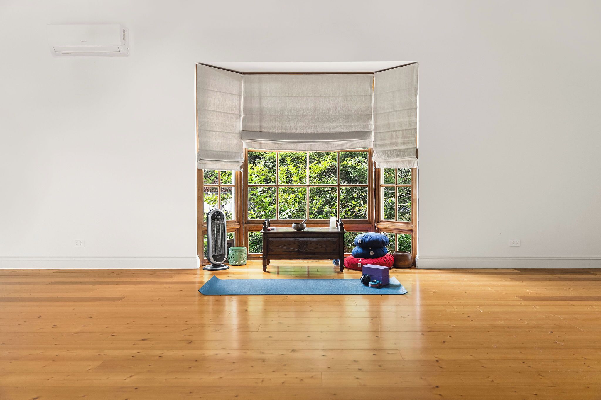 Una habitación con piso de madera, ventana con vista a follaje, un escritorio con silla y una esterilla de yoga.