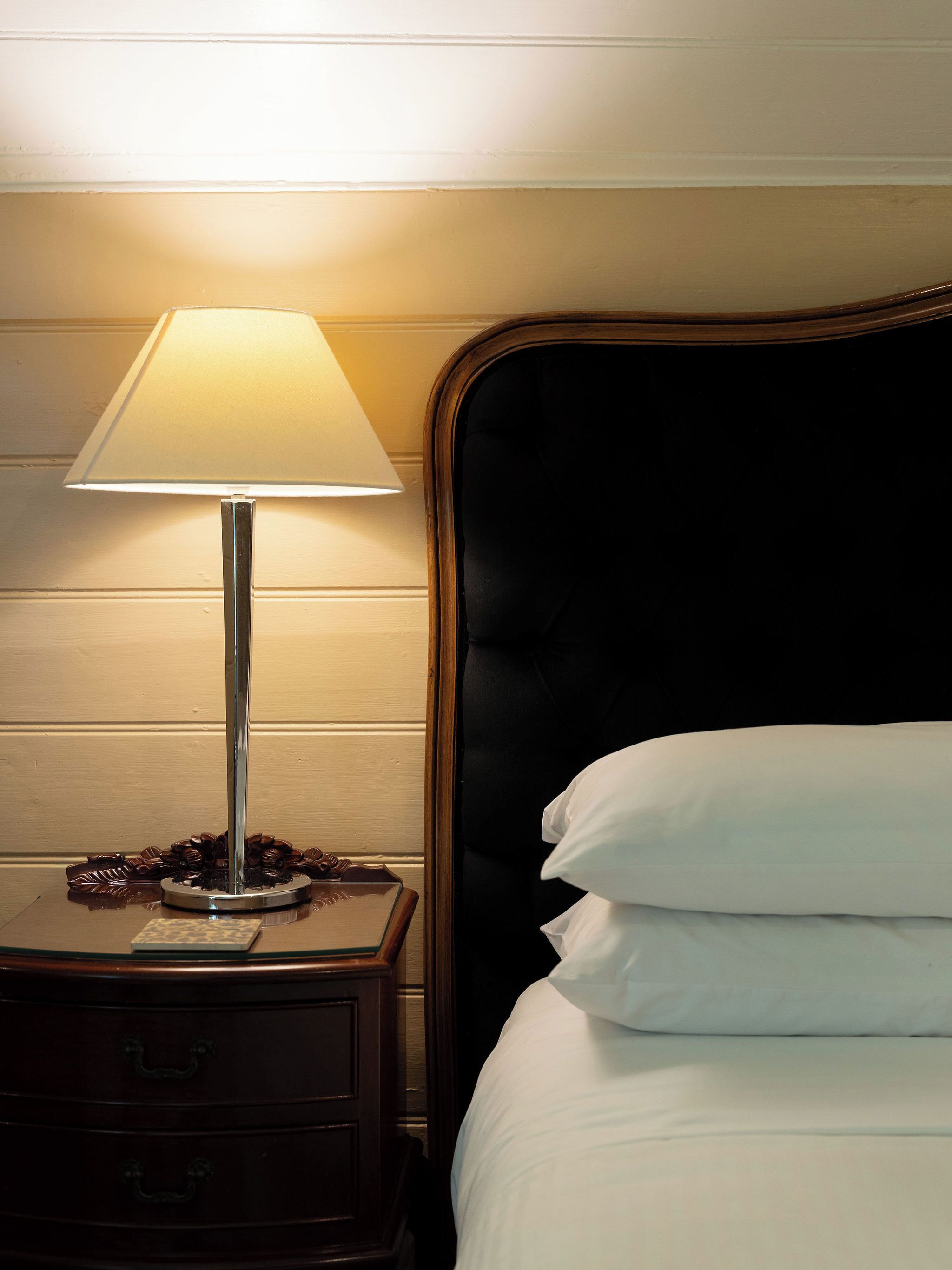 A bed with white pillows, a bedside lamp, a small table with a decorative item, and a wooden nightstand.
