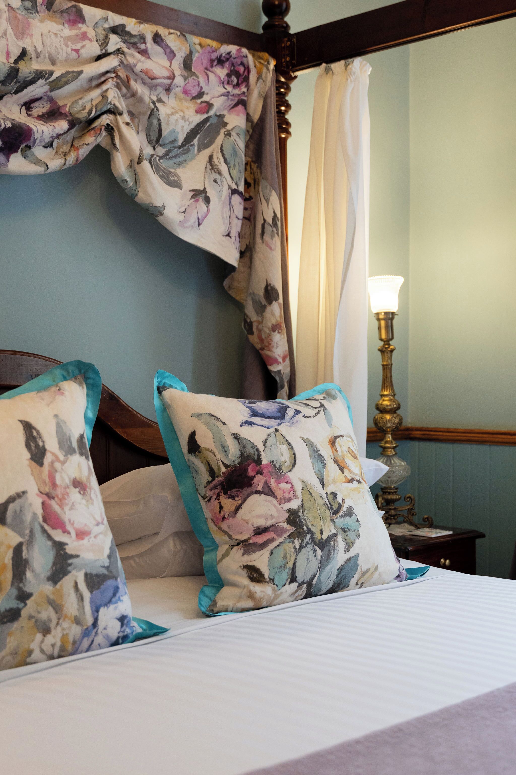 A bedroom with a canopy bed, floral-patterned pillows, a bedside table with a lamp, and a mirror.