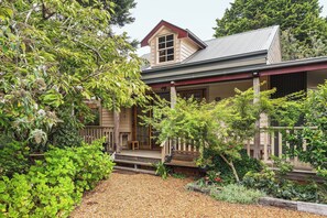 A house with a porch, surrounded by lush greenery and a gravel path. - Kurrara Historic Guest House (Katoomba)