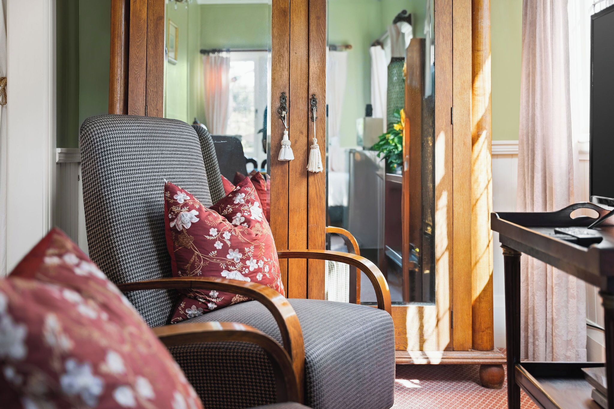 A room with a houndstooth patterned armchair, a wooden sideboard, and a mirror reflecting a green wall and a window.