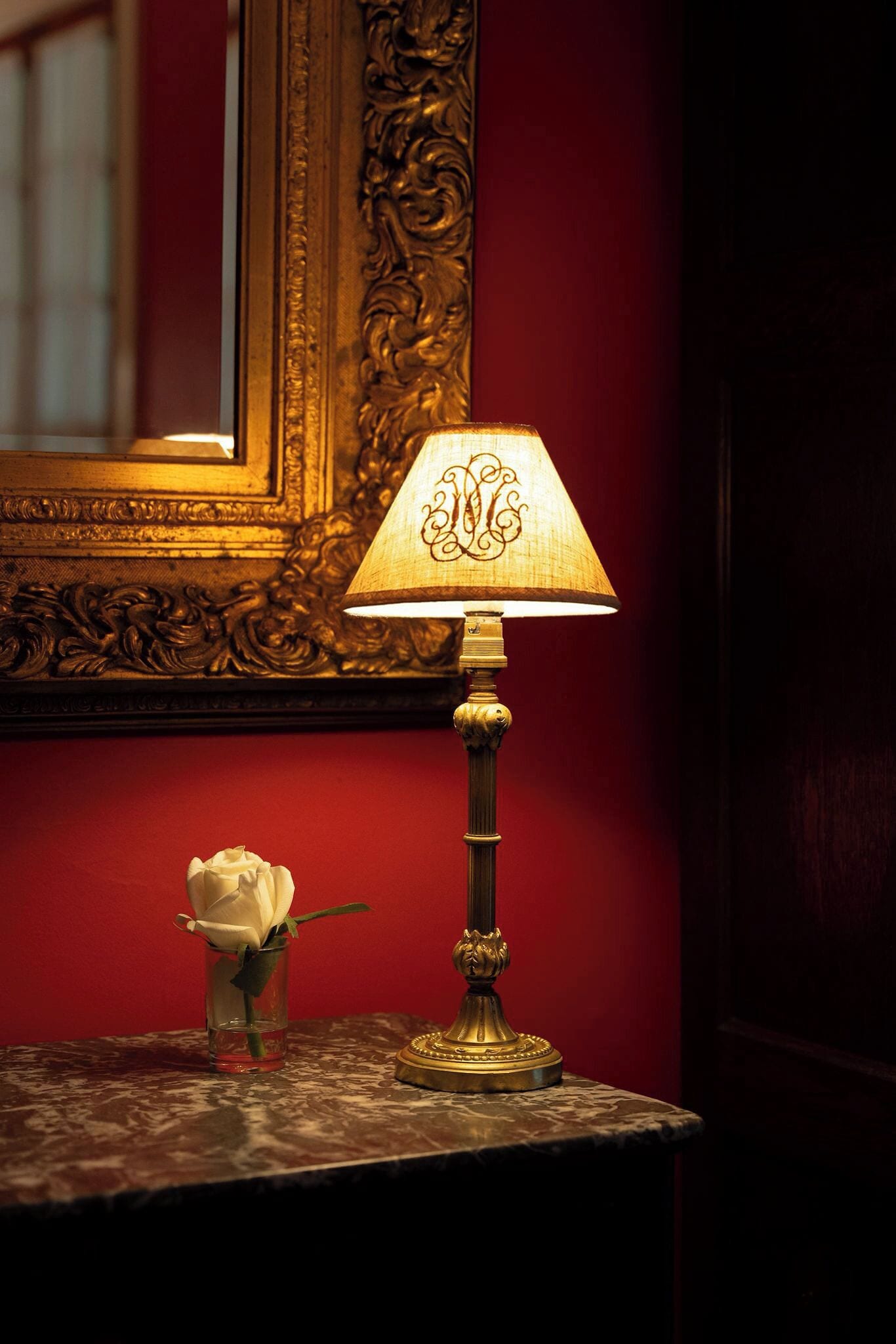 A lamp with a decorative shade, a vase with a single white rose, and a mirror with an ornate frame.