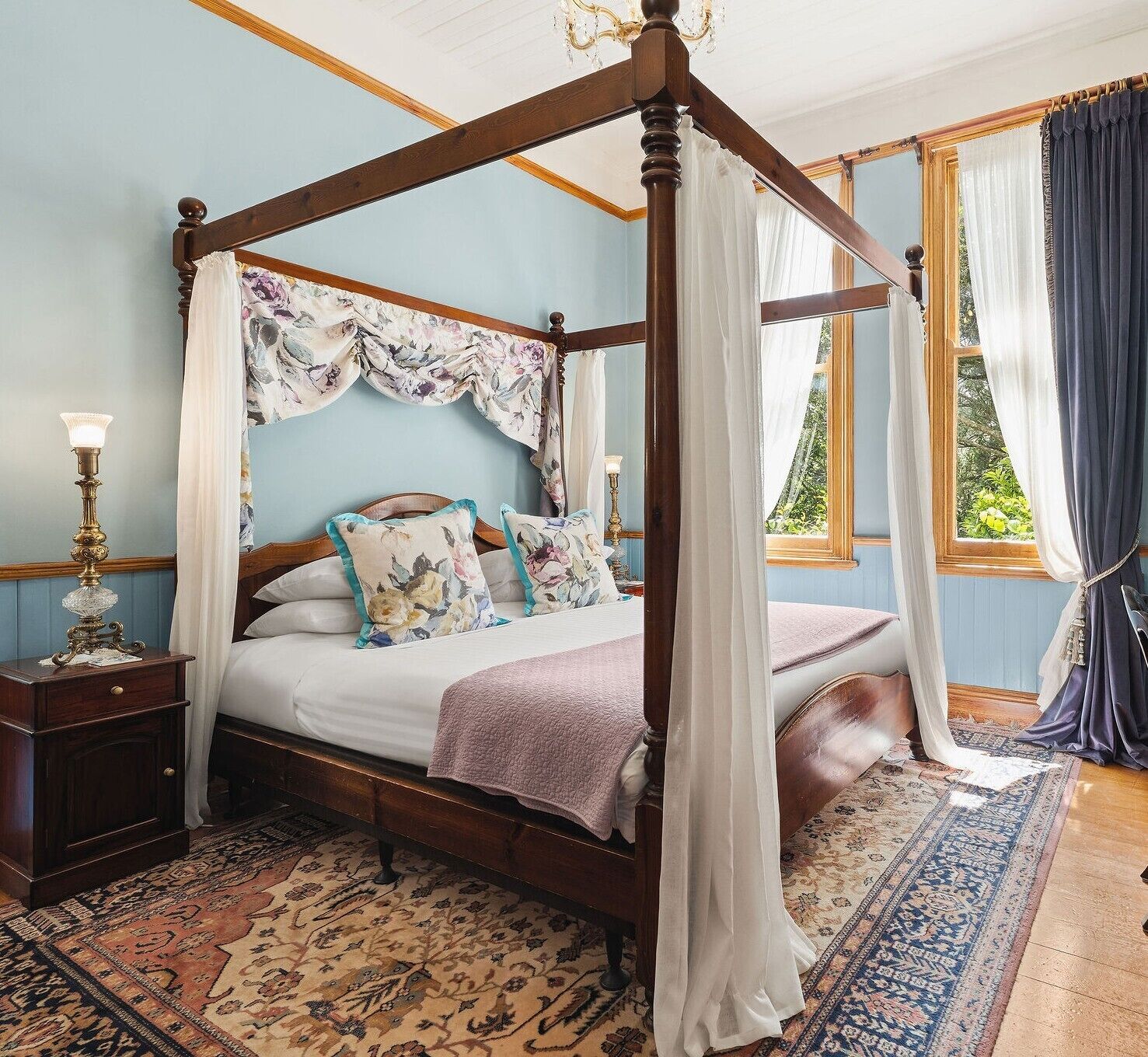 Ein Himmelsbett mit floraler Bettwäsche, ein Nachttisch mit Lampe und ein gemustertes Teppich.