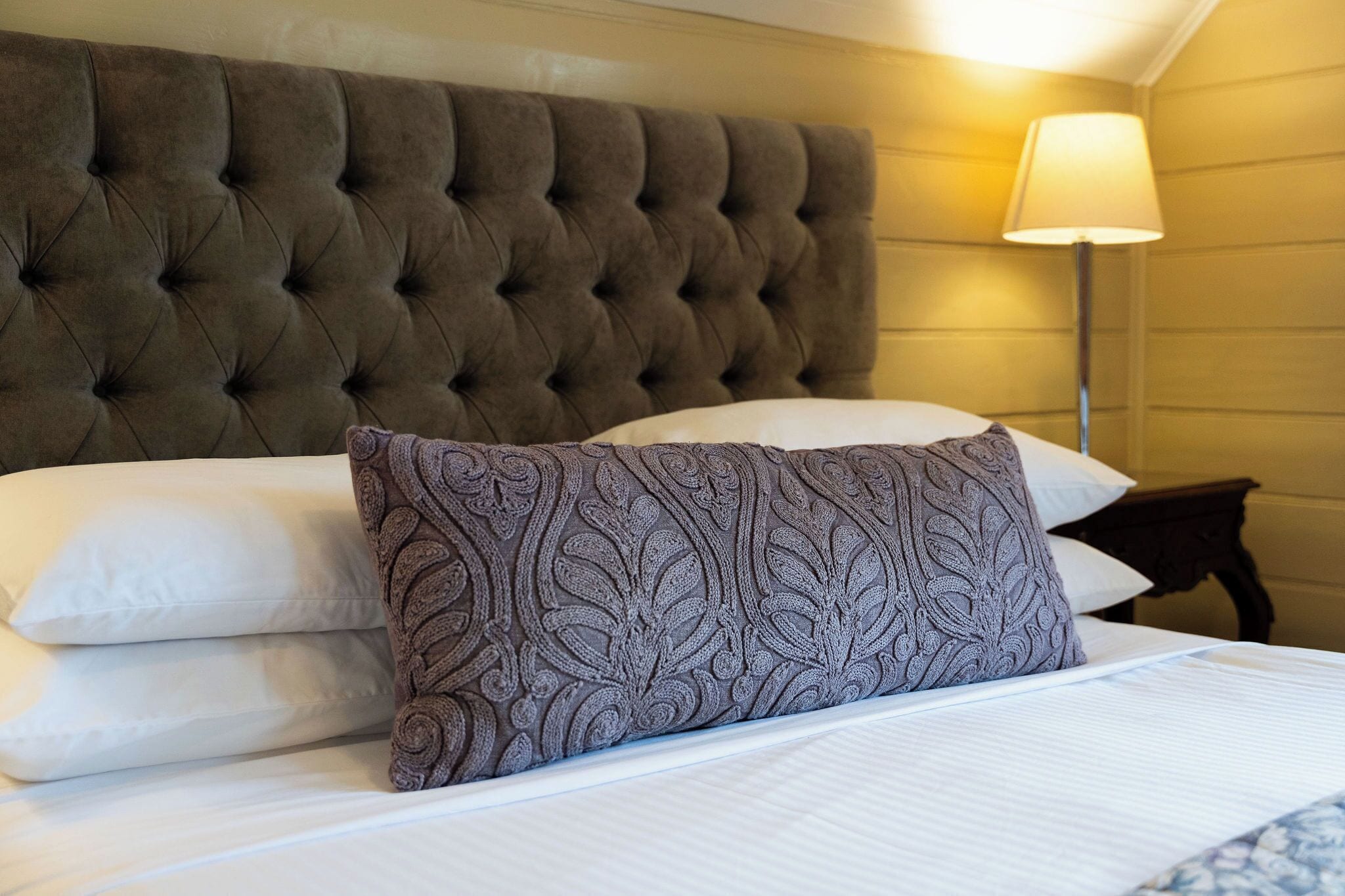 A neatly made bed with a tufted headboard, a patterned pillow, and a bedside lamp.