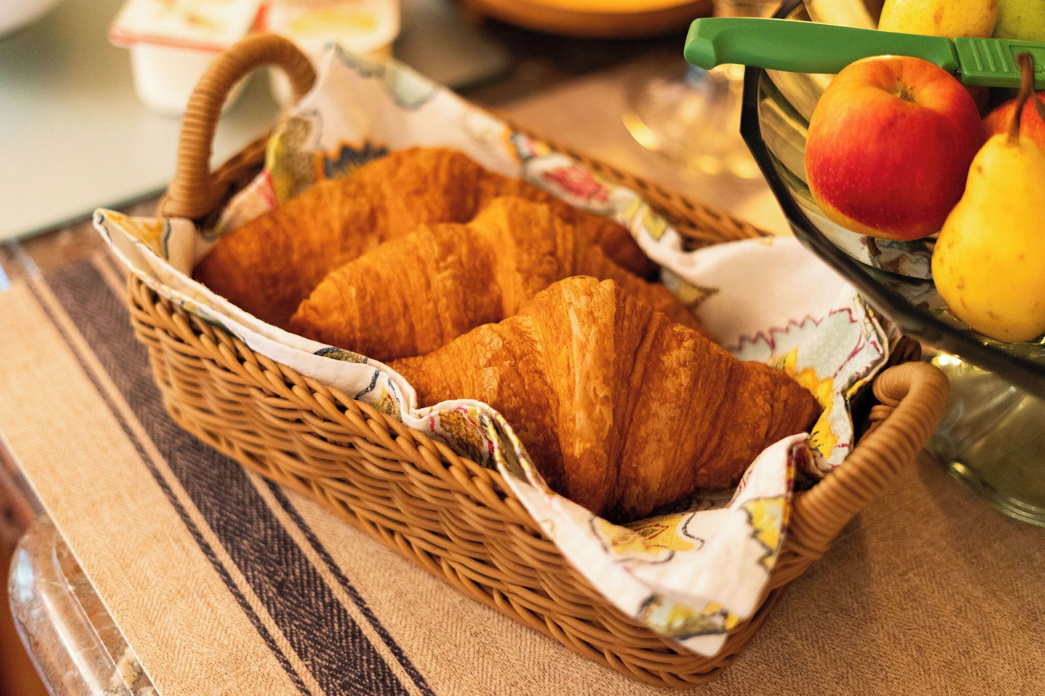 Una cesta de mimbre con cruasanes y un bol de fruta sobre una mesa.
