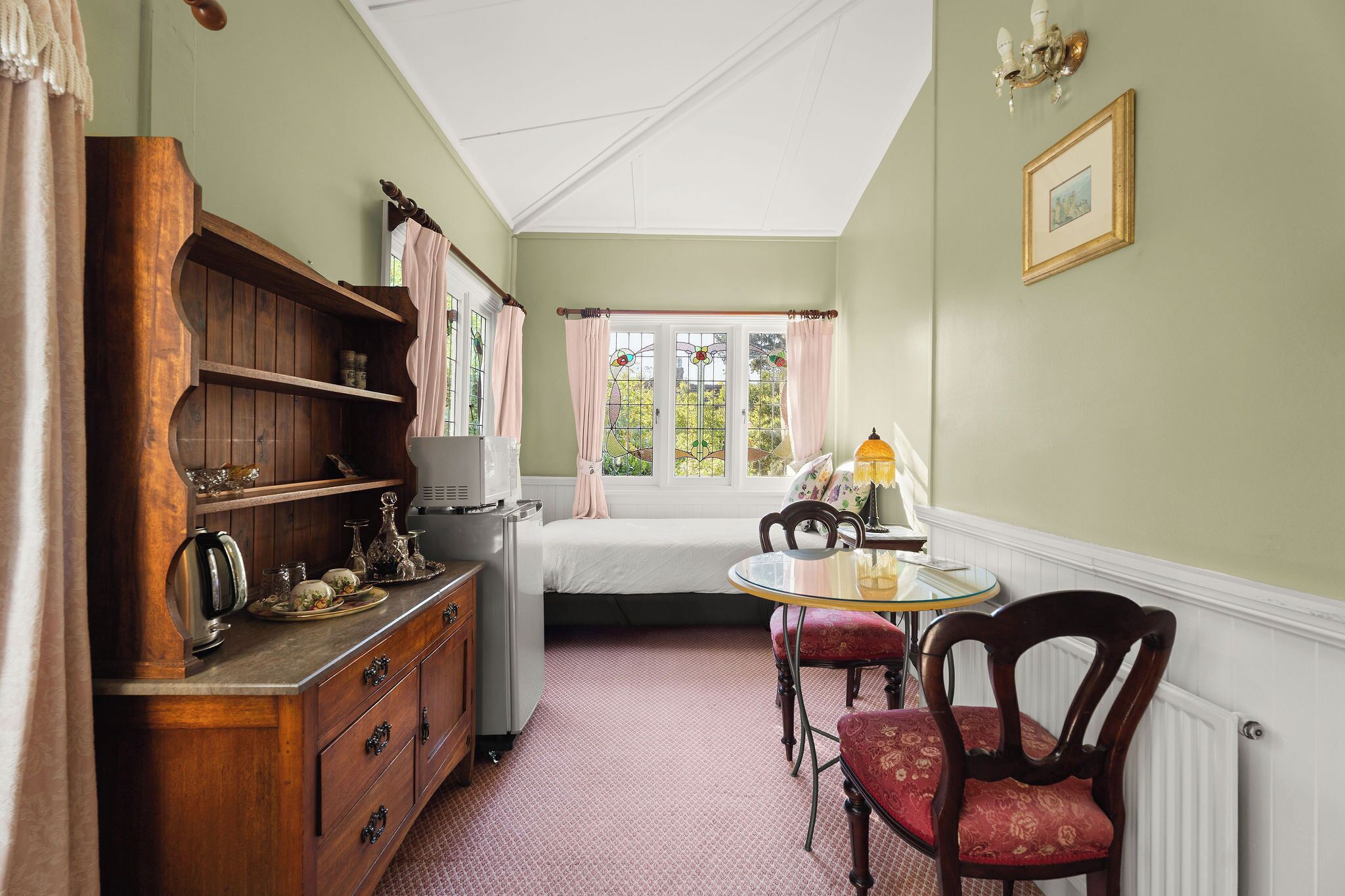 Ein Zimmer mit einem Bett, einem Holzschrank, einem Tisch mit Stühlen, einem Fenster mit Vorhängen und einer Chandelier.