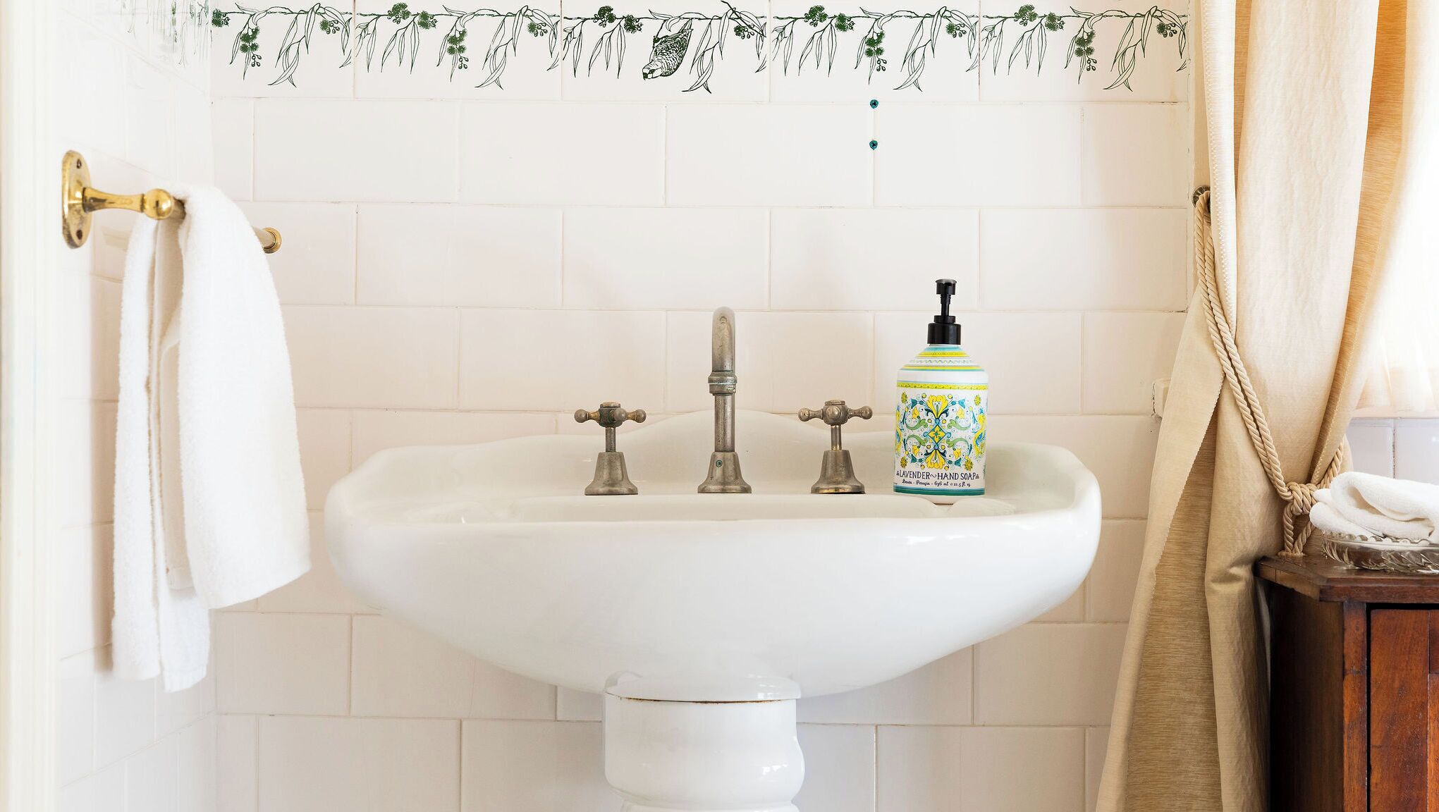 Une salle de bain avec un lavabo blanc, trois robinets, un flacon de savon liquide à la lavande et une serviette suspendue à un porte-serviettes en laiton.