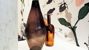 Un jarrón y una botella sobre una mesa, con una pared decorada con ilustraciones botánicas.
