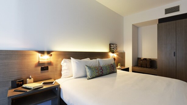 Una habitación de hotel con una cama grande, mesitas de noche, un armario y luces fijadas en la pared.
