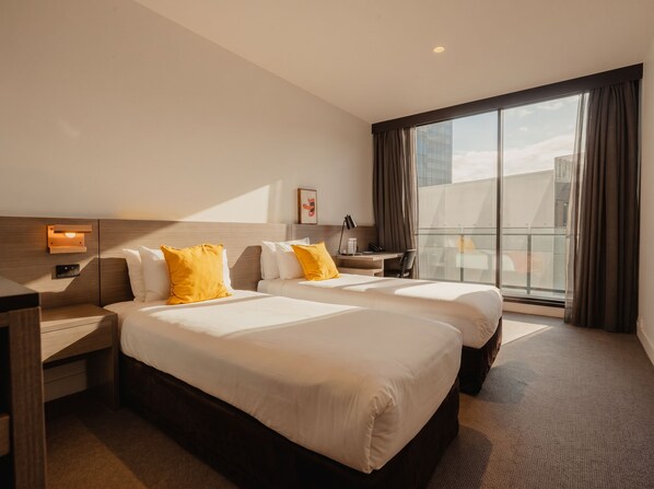 A hotel room with two beds, a desk, a chair, a nightstand, a lamp, and a large window with curtains.