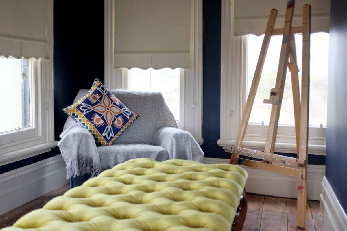 Une pièce comprenant un fauteuil gris, un coussin coloré, un pouf jaune à motifs et un chevalet en bois placé près d’une fenêtre.