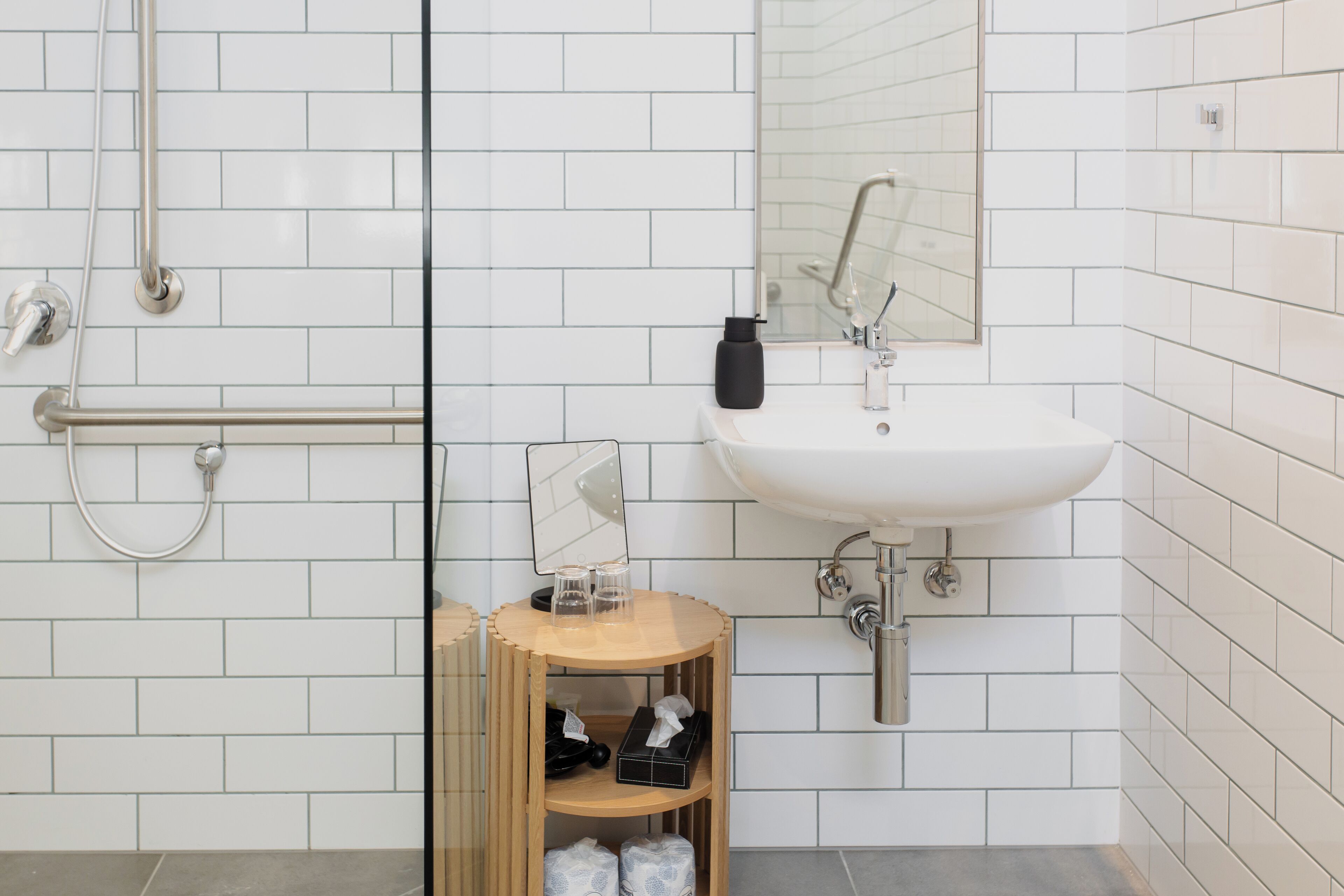 A modern bathroom with white tiled walls, a white sink, a mirror, and a wooden shelf with toiletries.