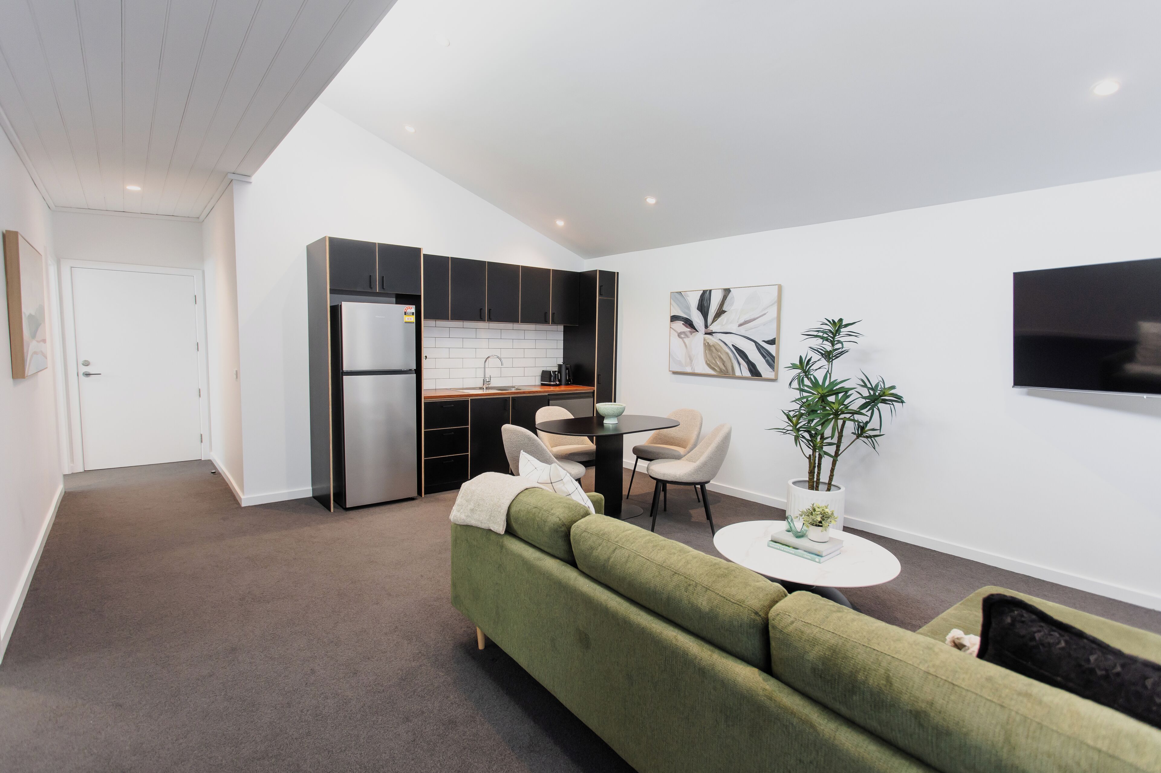A modern kitchen with a built-in refrigerator, a dining area with a round table and chairs, and a living room with a green sofa and a small coffee table.