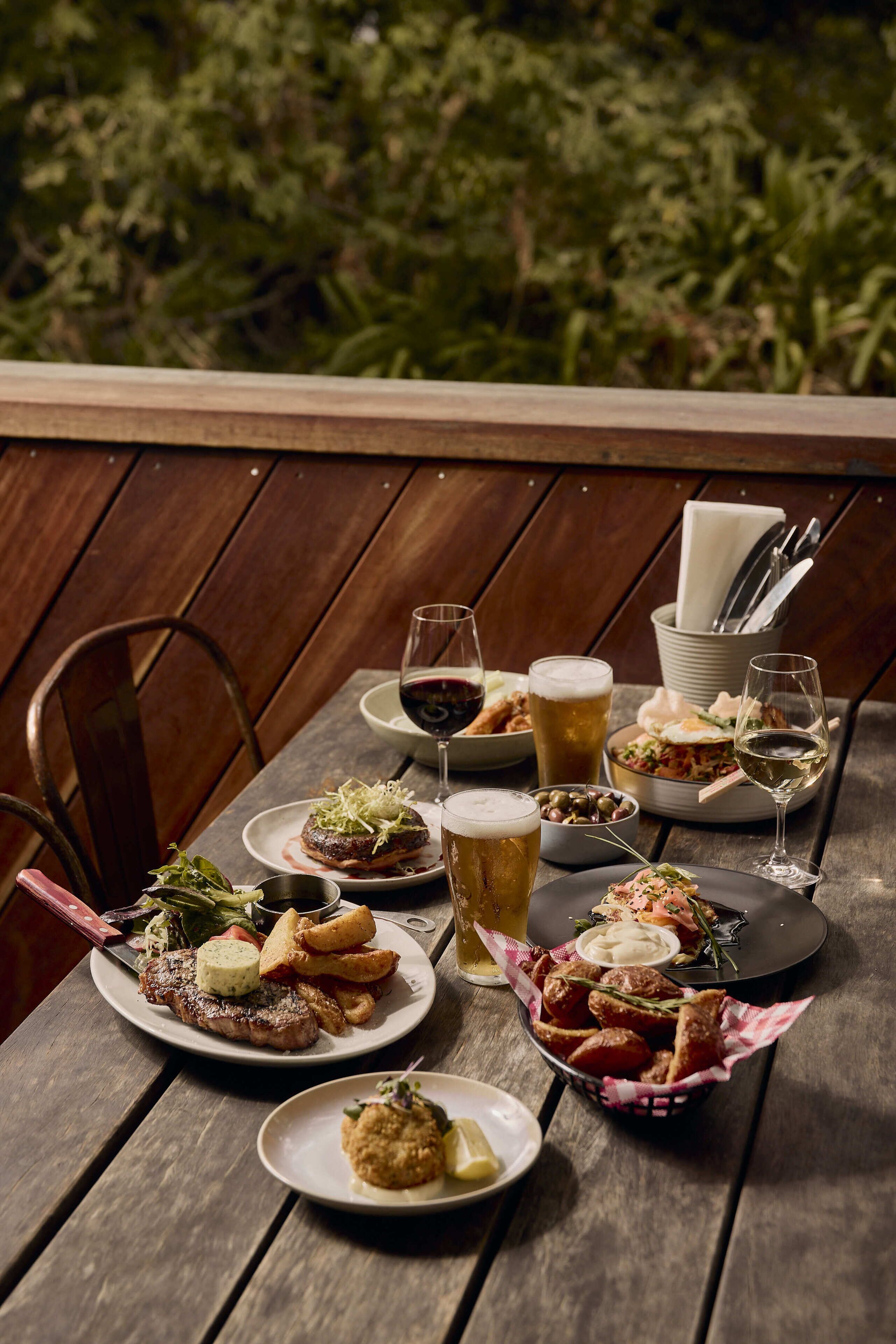 A wooden table set with various dishes, including steak, fries, and salad, along with multiple glasses of beer and wine.