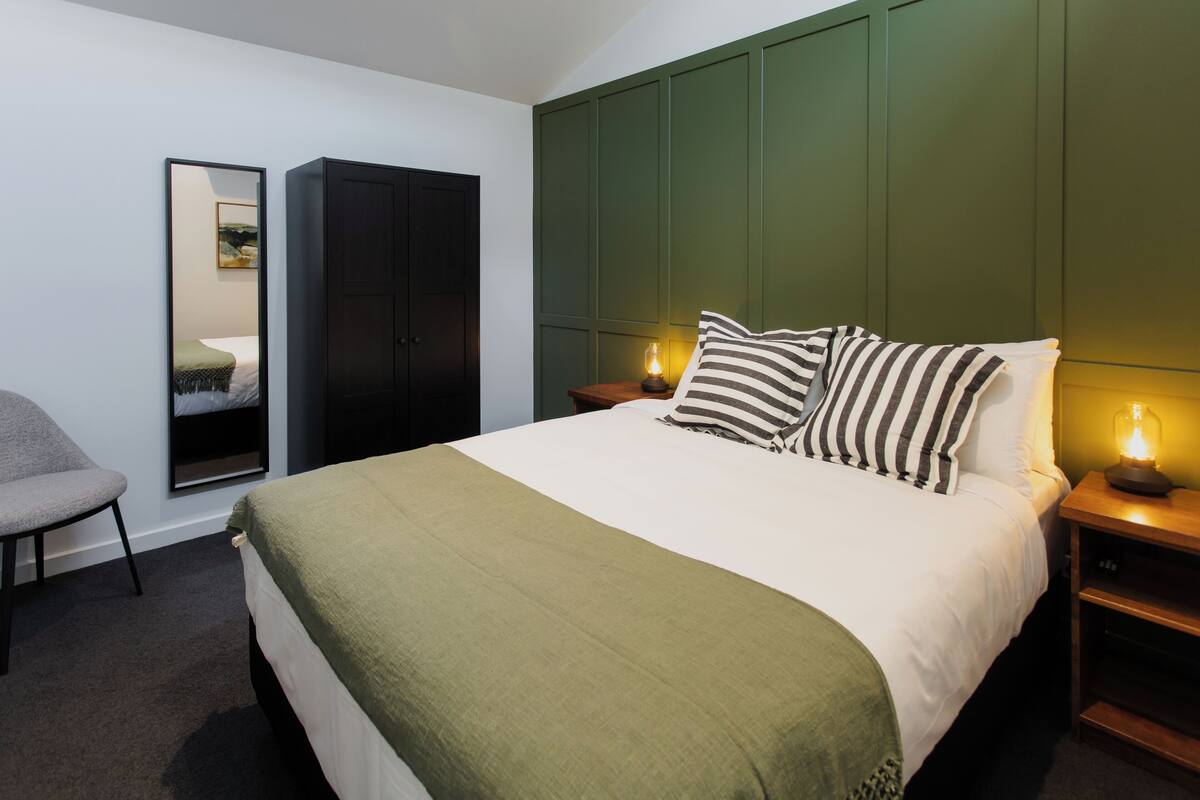Une chambre bien rangée, avec un lit, une chaise, une armoire et une table de chevet sur laquelle se trouve une lampe.