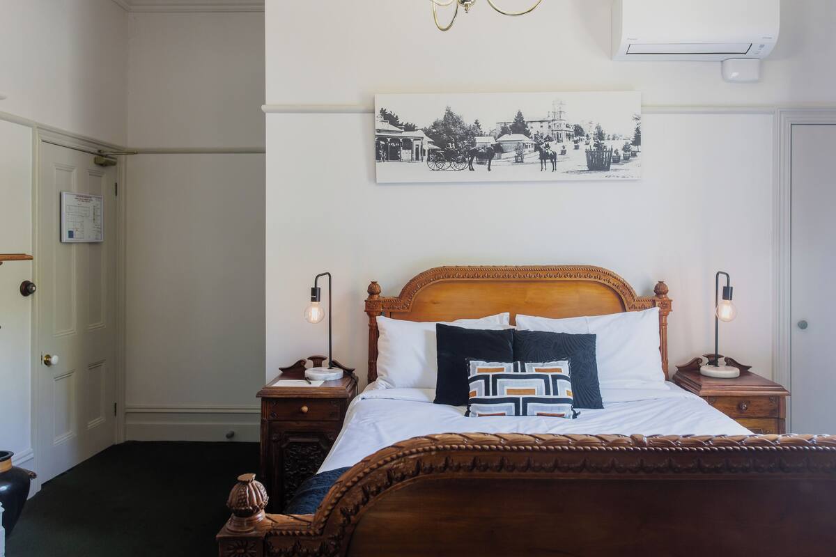 Une chambre à coucher bien rangée, avec un lit en bois, des tables de chevet, un climatiseur fixé au mur et un tableau encadré au-dessus du lit.