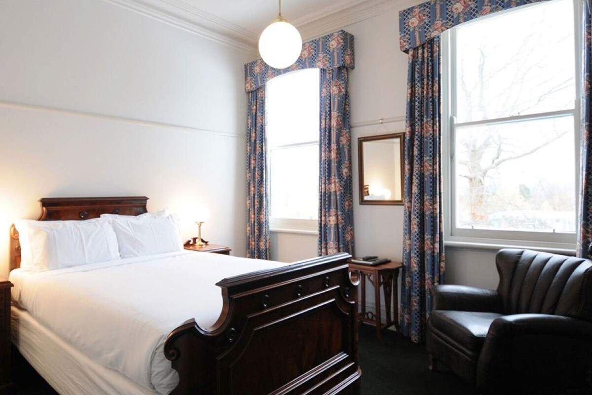 A hotel room with a large bed, a chair, a small table, and a window with curtains.