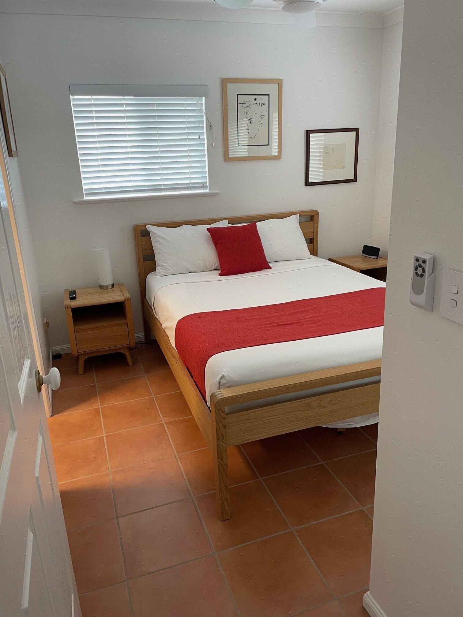 Een netjes ingerichte slaapkamer met een houten bed, wit beddengoed, een rode plaid, een nachtkastje met een lamp en een raam met jaloezieën.