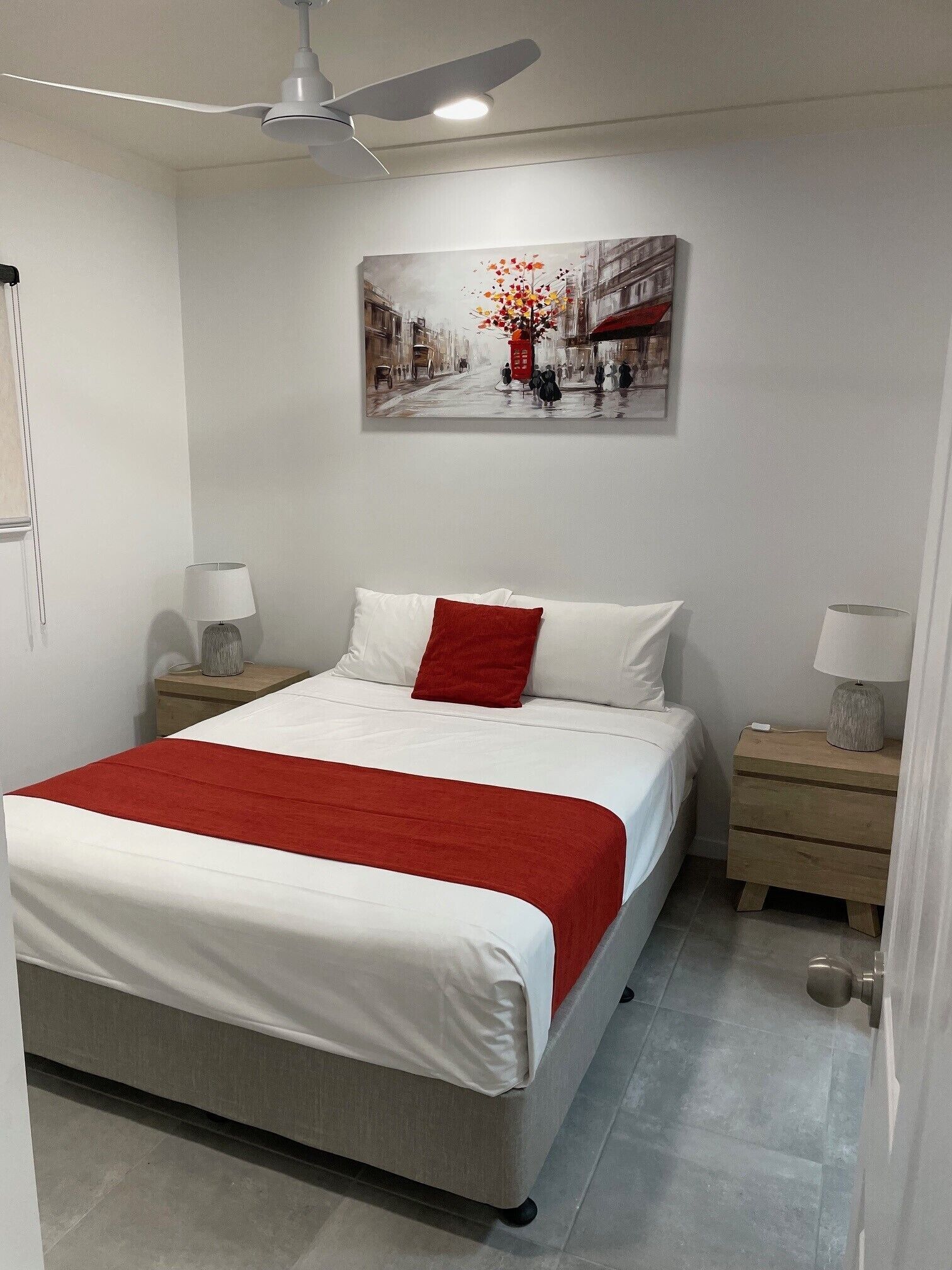 Ein Schlafzimmer mit einem Bett, einer roten Tagesdecke, einem Gemälde einer Straßenszene und einem Deckenventilator.