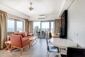 A modern living room with a sofa, coffee table, and dining area, large windows with curtains, and a view of the city. - Oaks Darwin Elan Hotel (Darwin)