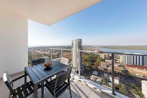 Un balcon avec une table et des chaises, donnant sur un paysage urbain avec des bâtiments et une étendue d’eau.