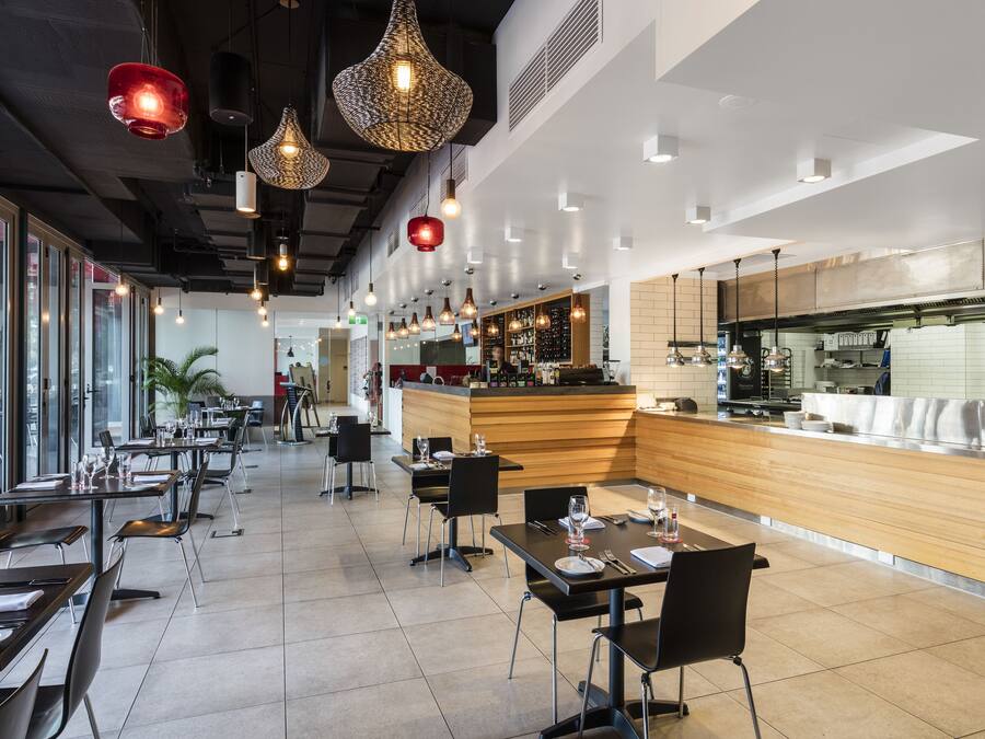 A modern restaurant with multiple dining tables and chairs, a bar area, and large windows allowing natural light.