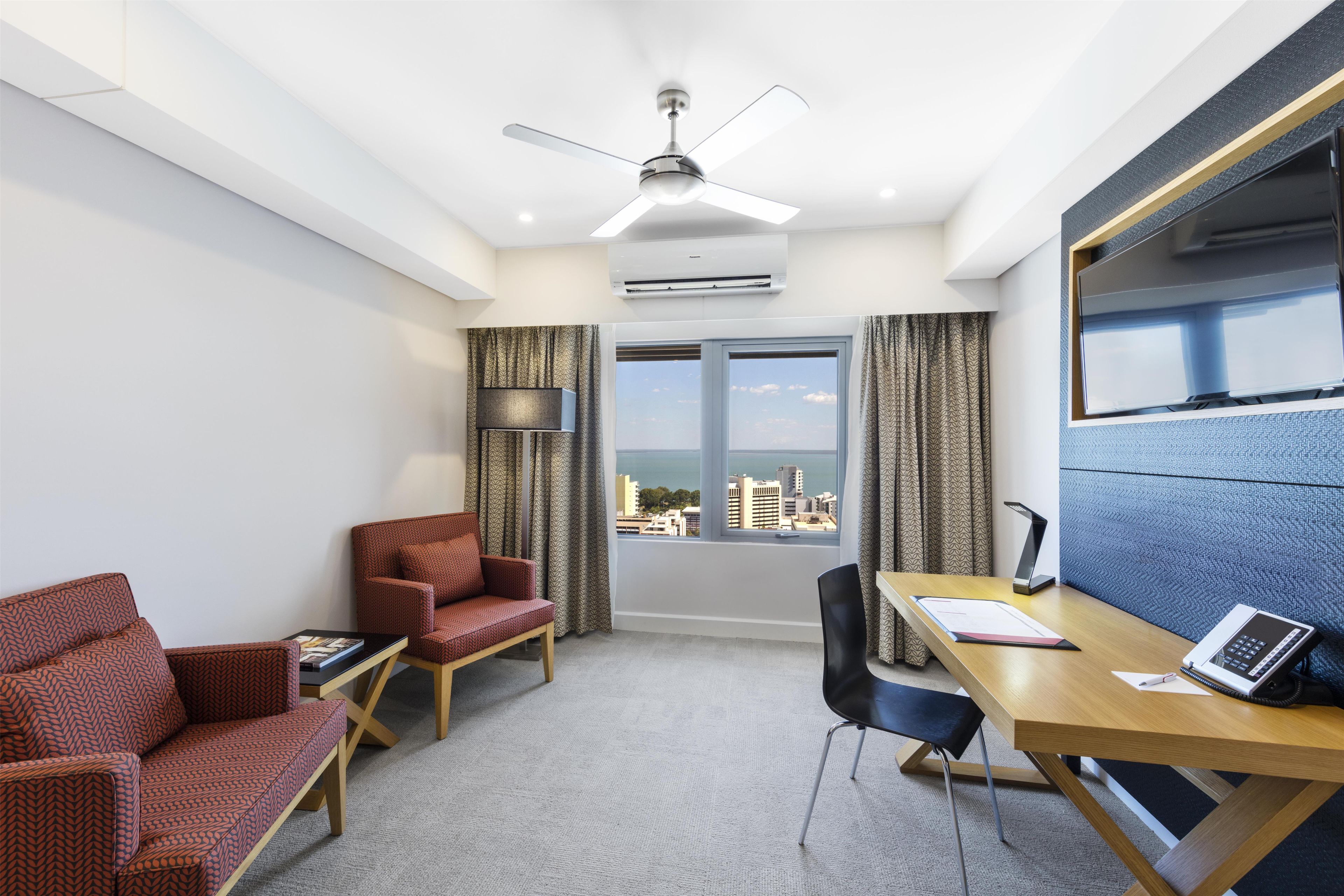 Una habitación de hotel con escritorio, dos sillas, ventilador de techo, equipo de aire acondicionado, ventana grande con cortinas y vista a la ciudad.
