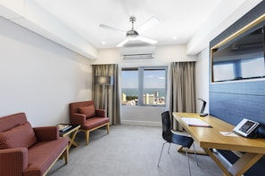 Una habitación de hotel con escritorio, dos sillas, ventilador de techo, equipo de aire acondicionado, ventana grande con cortinas y vista a la ciudad.