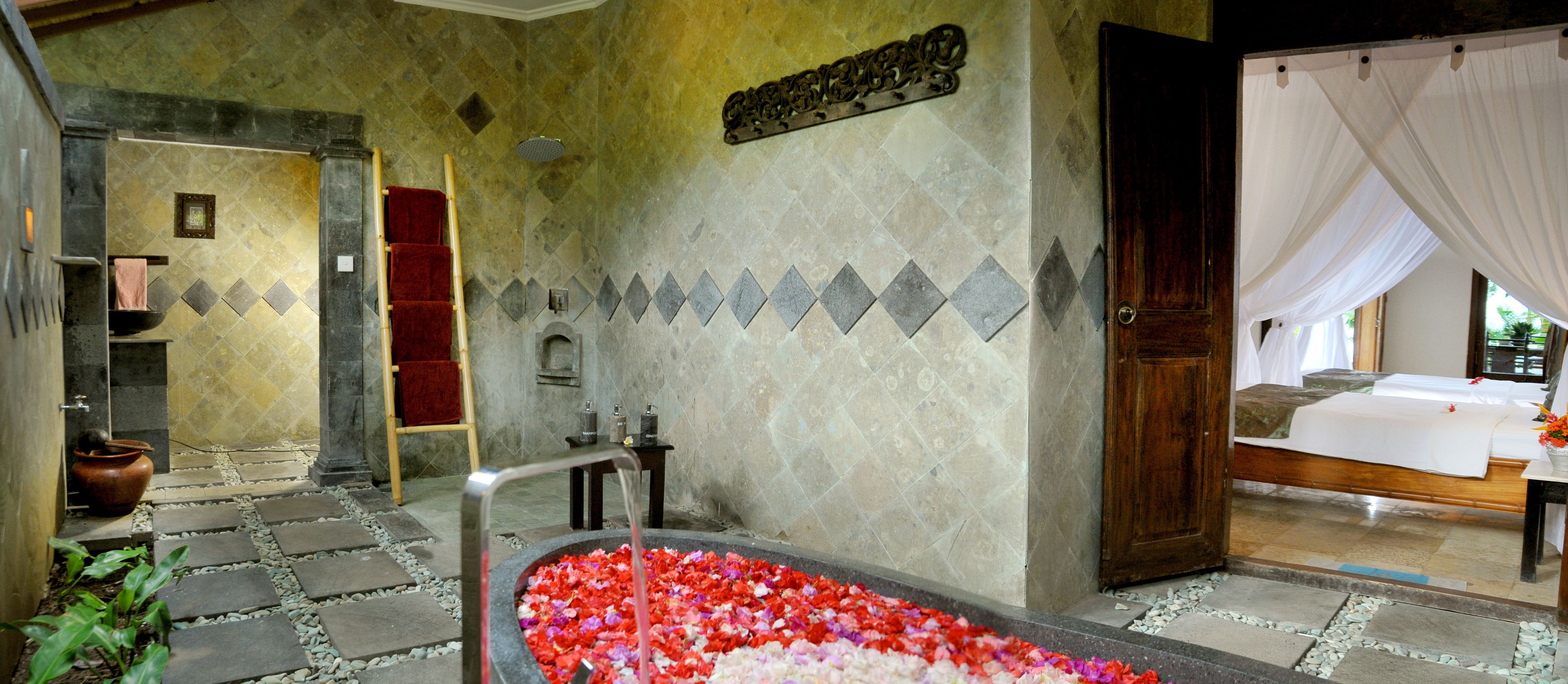 Un baño con suelo de piedra, una bañera grande llena de pétalos de rosa, una escalera de madera y una cama con dosel blanco en la habitación contigua.