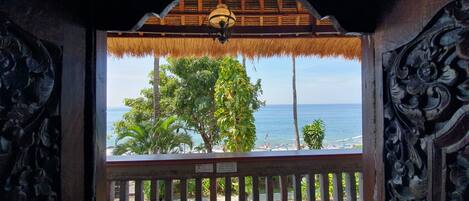 A view from a room with a thatched roof and ornate metal railings, overlooking a beach with palm trees and the ocean.