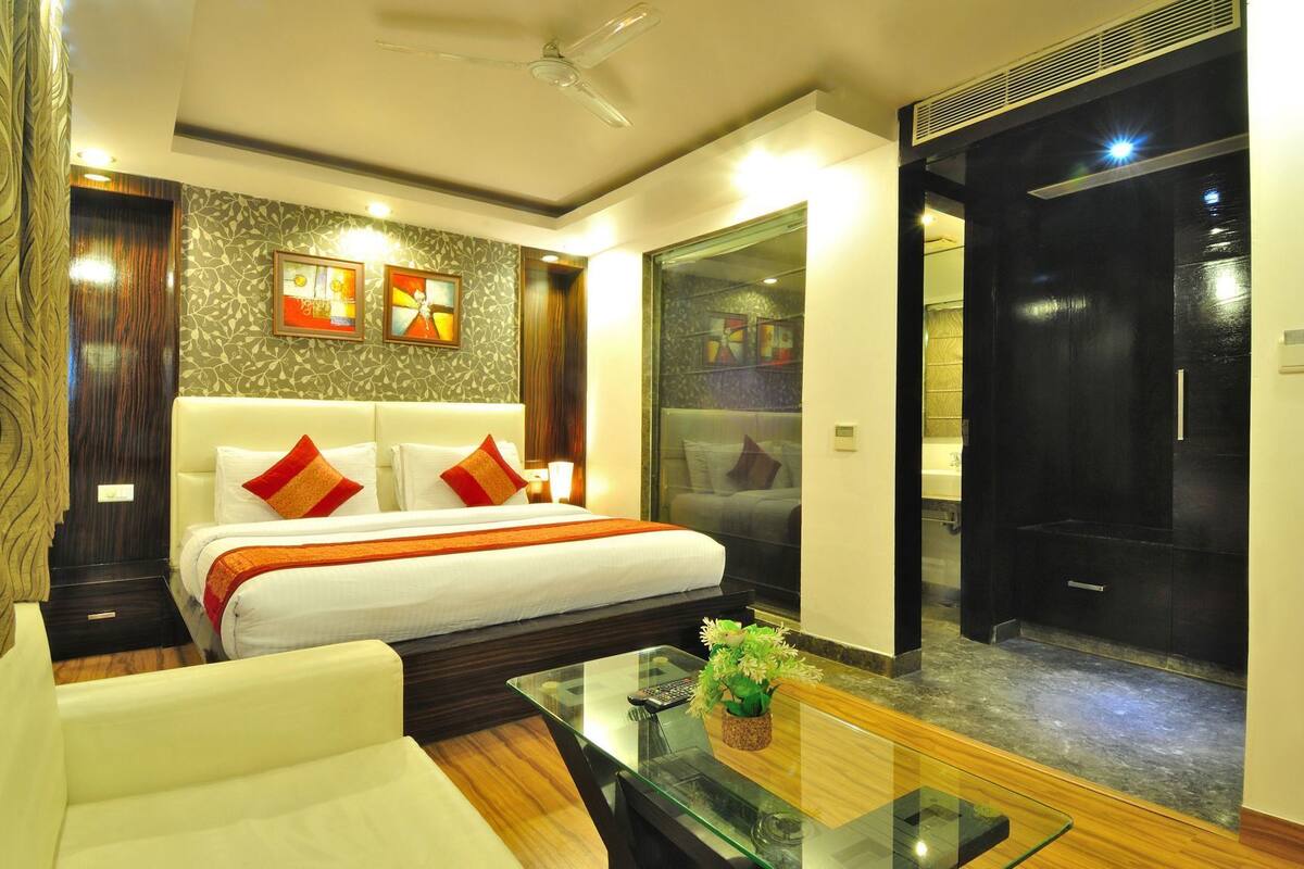 Habitación de hotel con cama, sofá, mesita de vidrio y baño visible a través de una puerta de cristal.