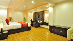 Una habitación de hotel moderna con piso de madera, cama con ropa de cama blanca y un detalle rojo, escritorio con silla, espejo grande y televisor montado en la pared.