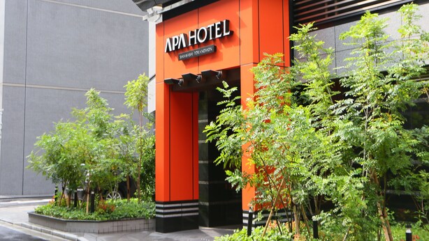The exterior of the APA Hotel with a prominent orange and black facade, surrounded by greenery and modern architecture. - APA Hotel Shimbashi Toranomon (Tokyo)